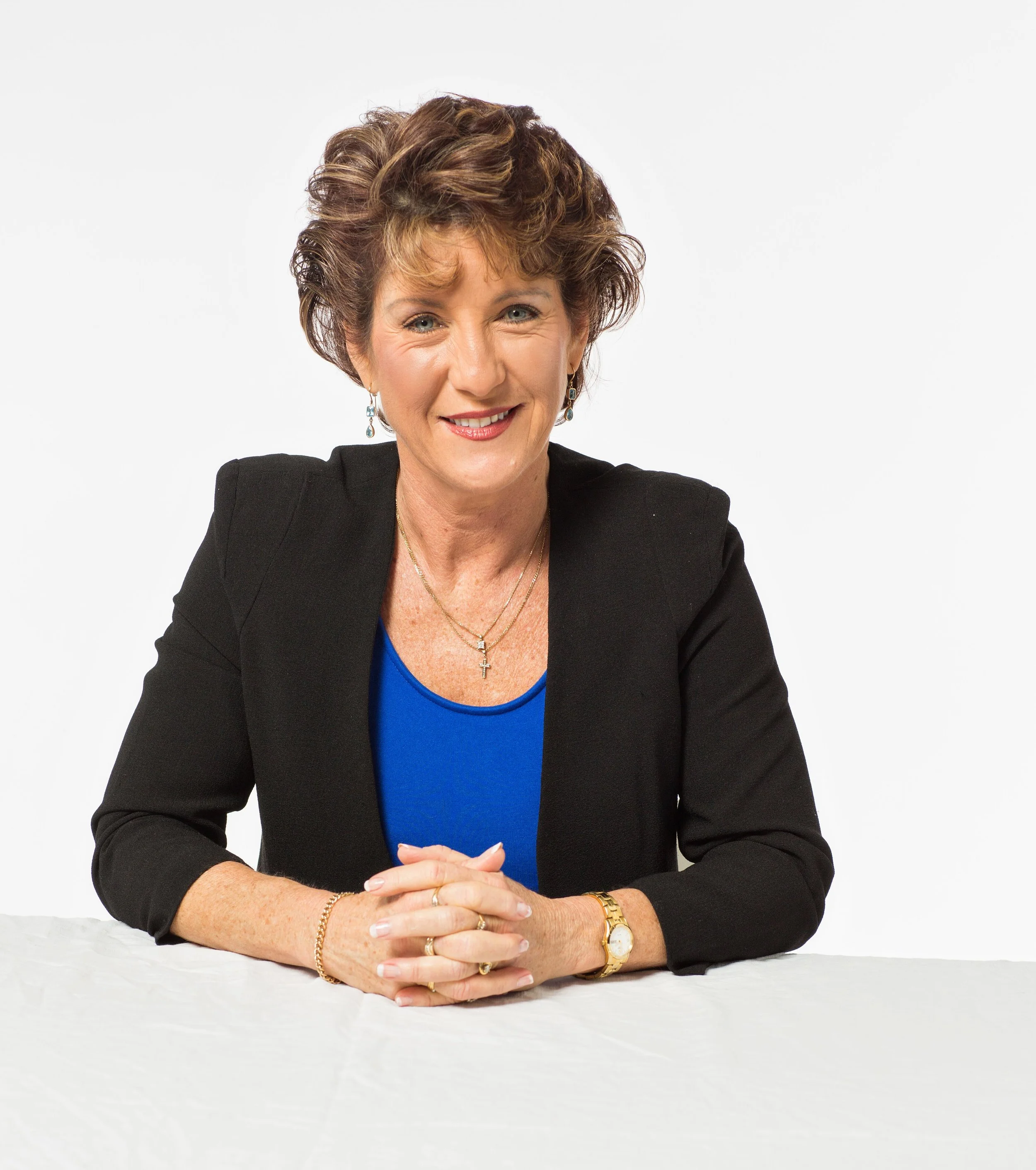 A smiling woman with short curly hair wearing a black blazer over a blue top, sitting at a table with her hands clasped.