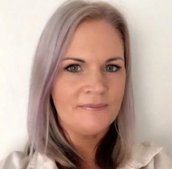 A woman with long, light purple hair and blue eyes looks at the camera against a plain white background.