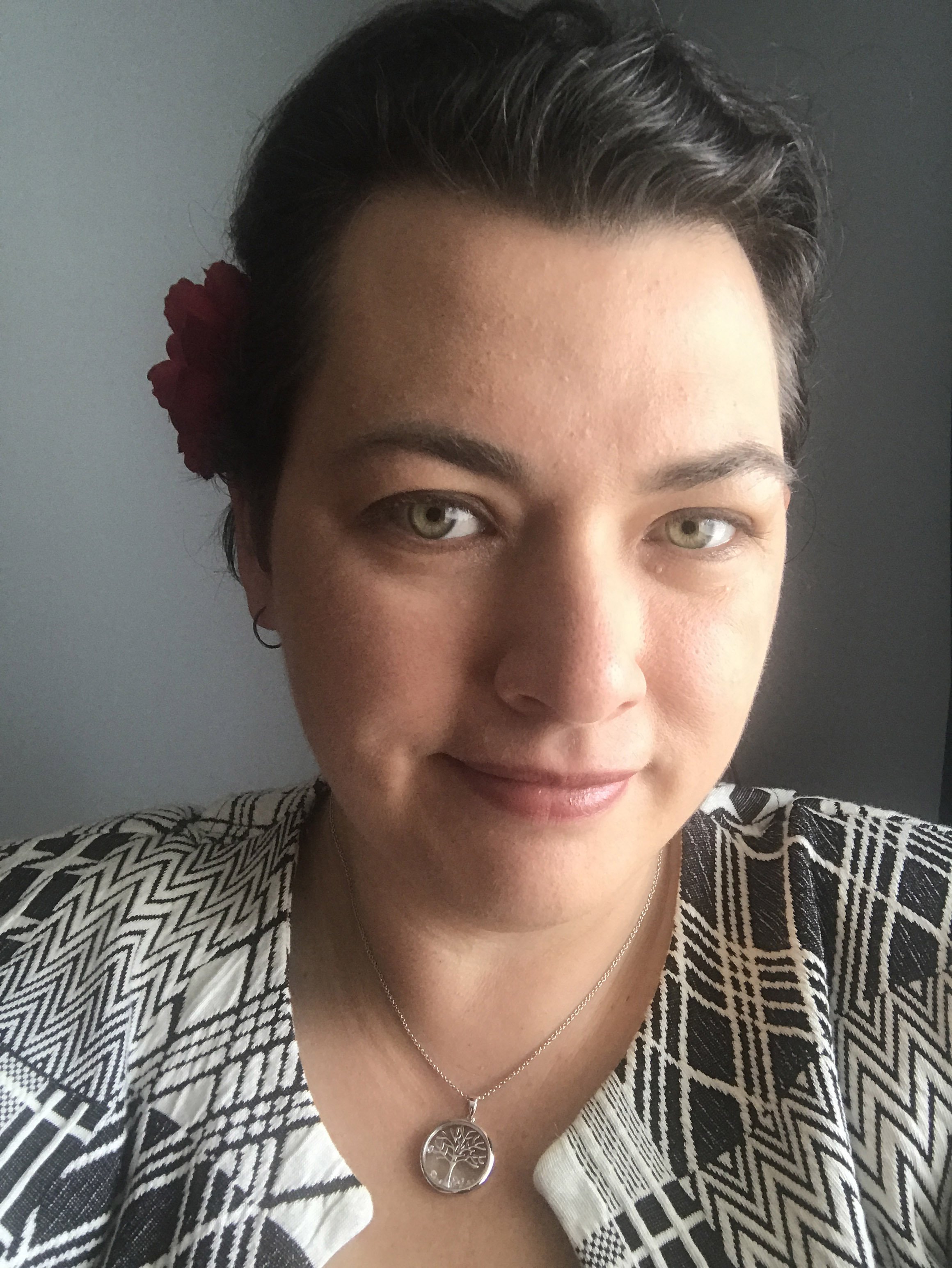 Close-up photo of a woman with short dark hair, light skin, wearing a patterned black and white shirt and a silver necklace with a tree pendant, standing against a dark wall with a red flower behind her ear.