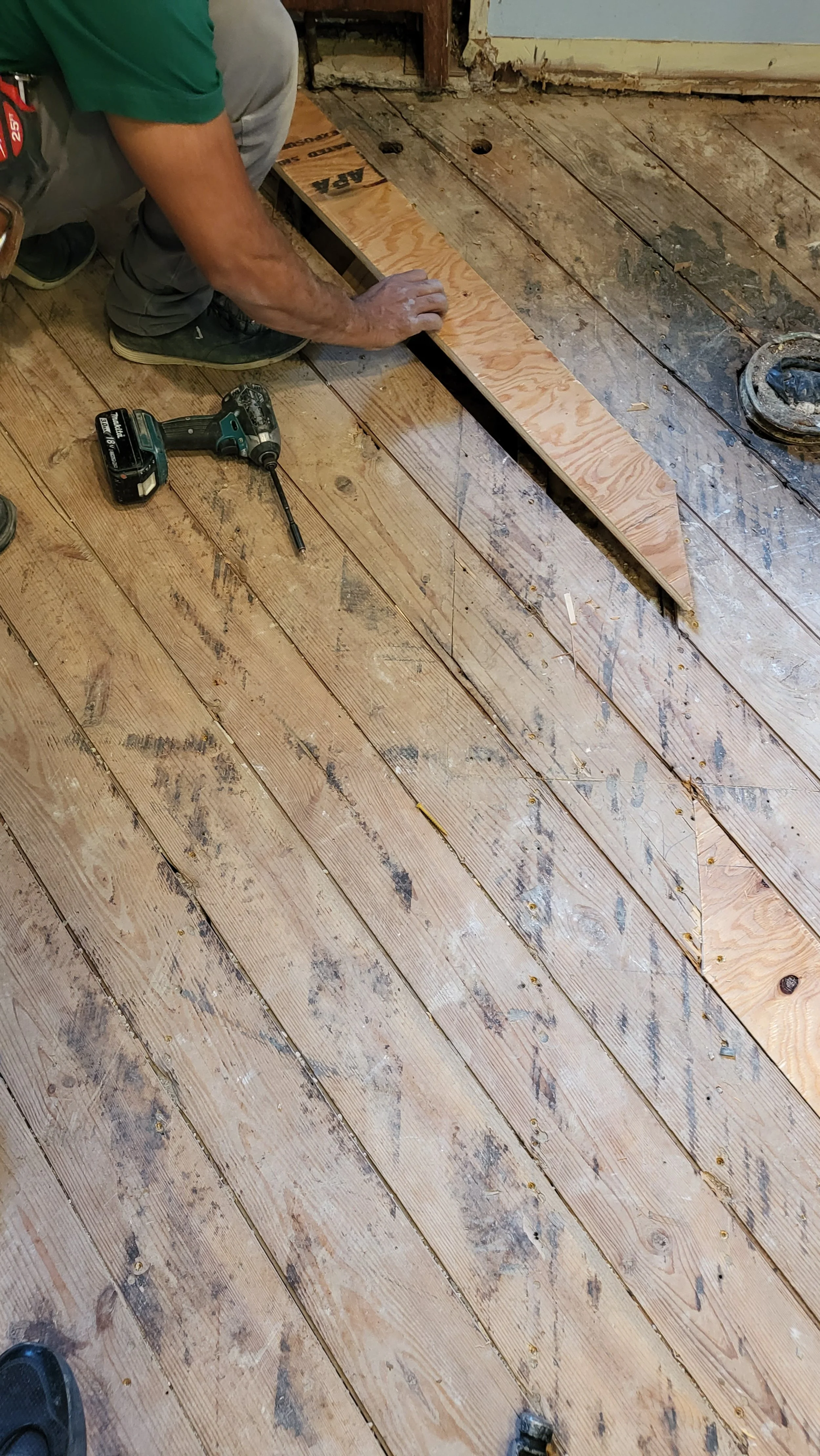 Person installing new wooden floorboards on a room with worn old wood flooring, with a power drill on the floor.