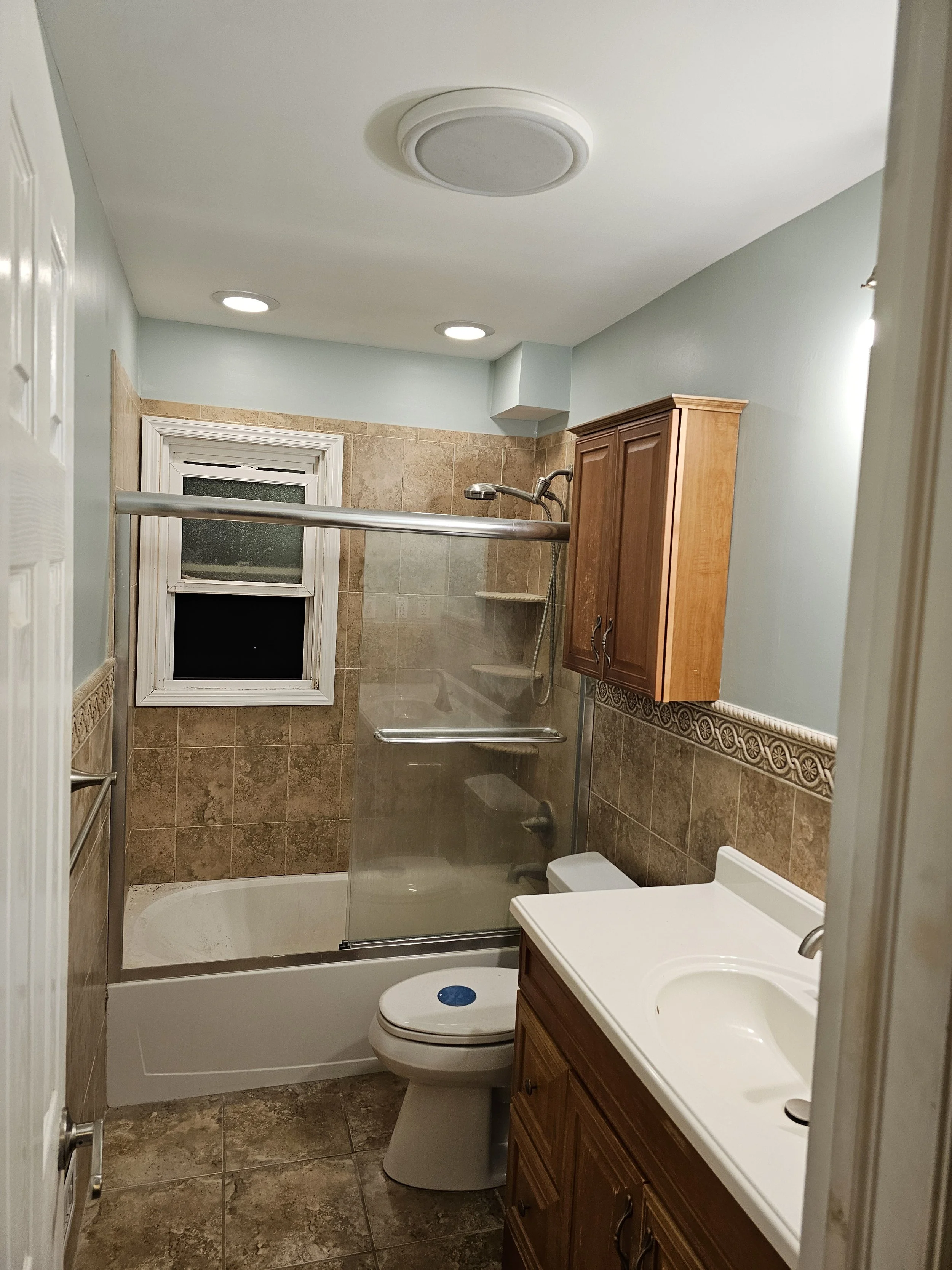 Small bathroom with beige tiled walls and floor, a white bathtub with a glass shower door, a window, a toilet, a wooden vanity with a sink, and a wooden cabinet above the toilet.