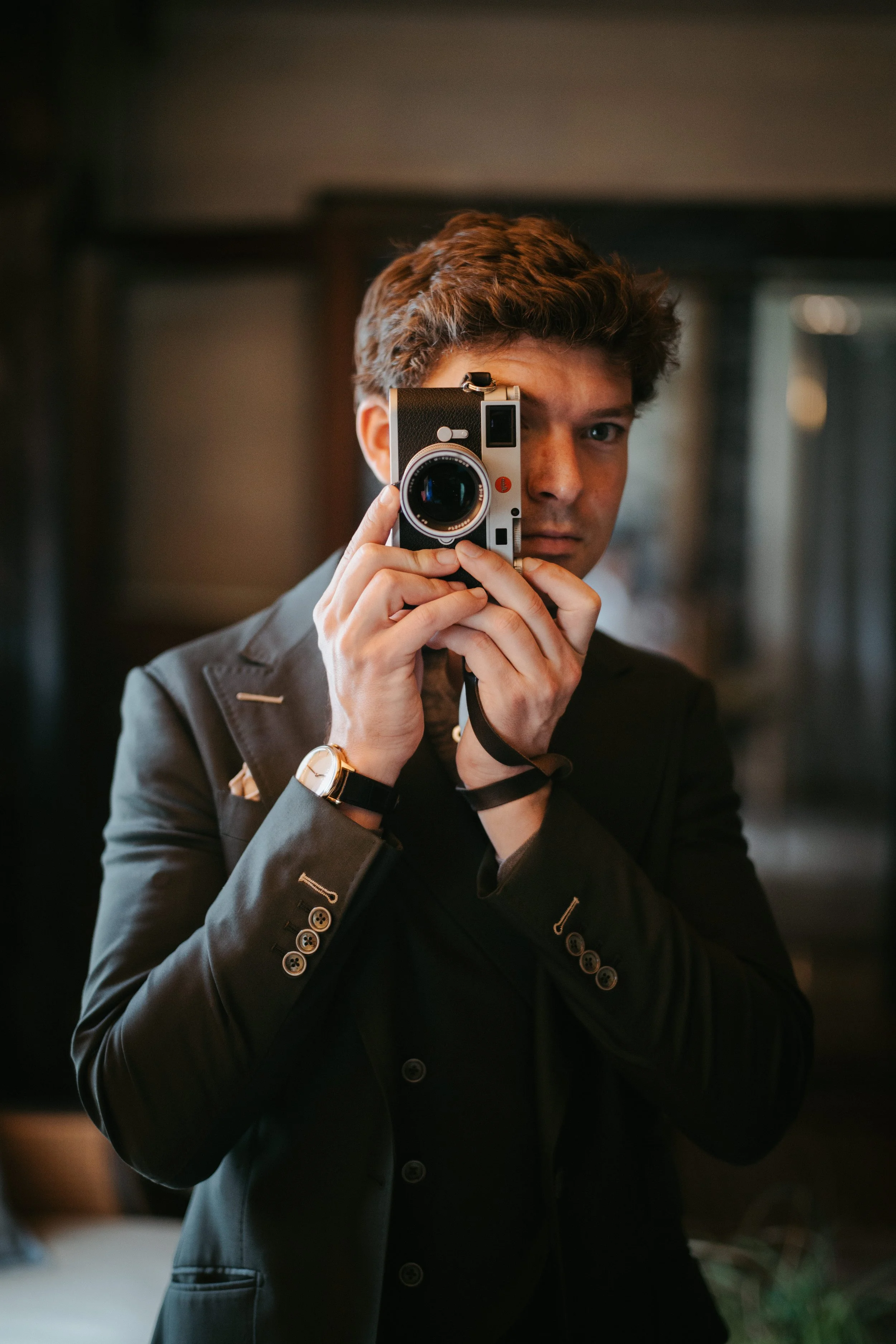 A smiling man with glasses and a beard wearing a blue blazer, holding a camera, outdoors with blurred trees and lights in the background.