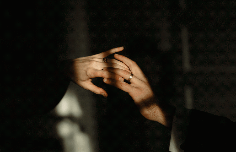 Two people gently holding hands, gently touching fingers, with focus on their rings, in a dimly lit setting.