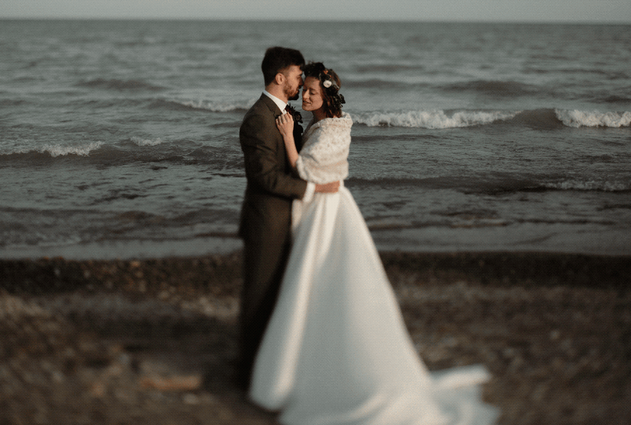 A bride and groom embrace on a beach, with water and small waves in the background.