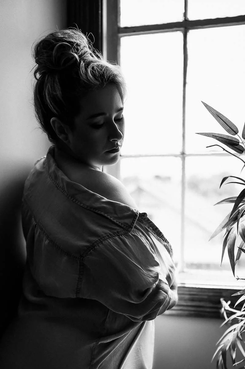 Boudoir portrait of woman in bun lit by the window and pulling her shirt off her shoulder