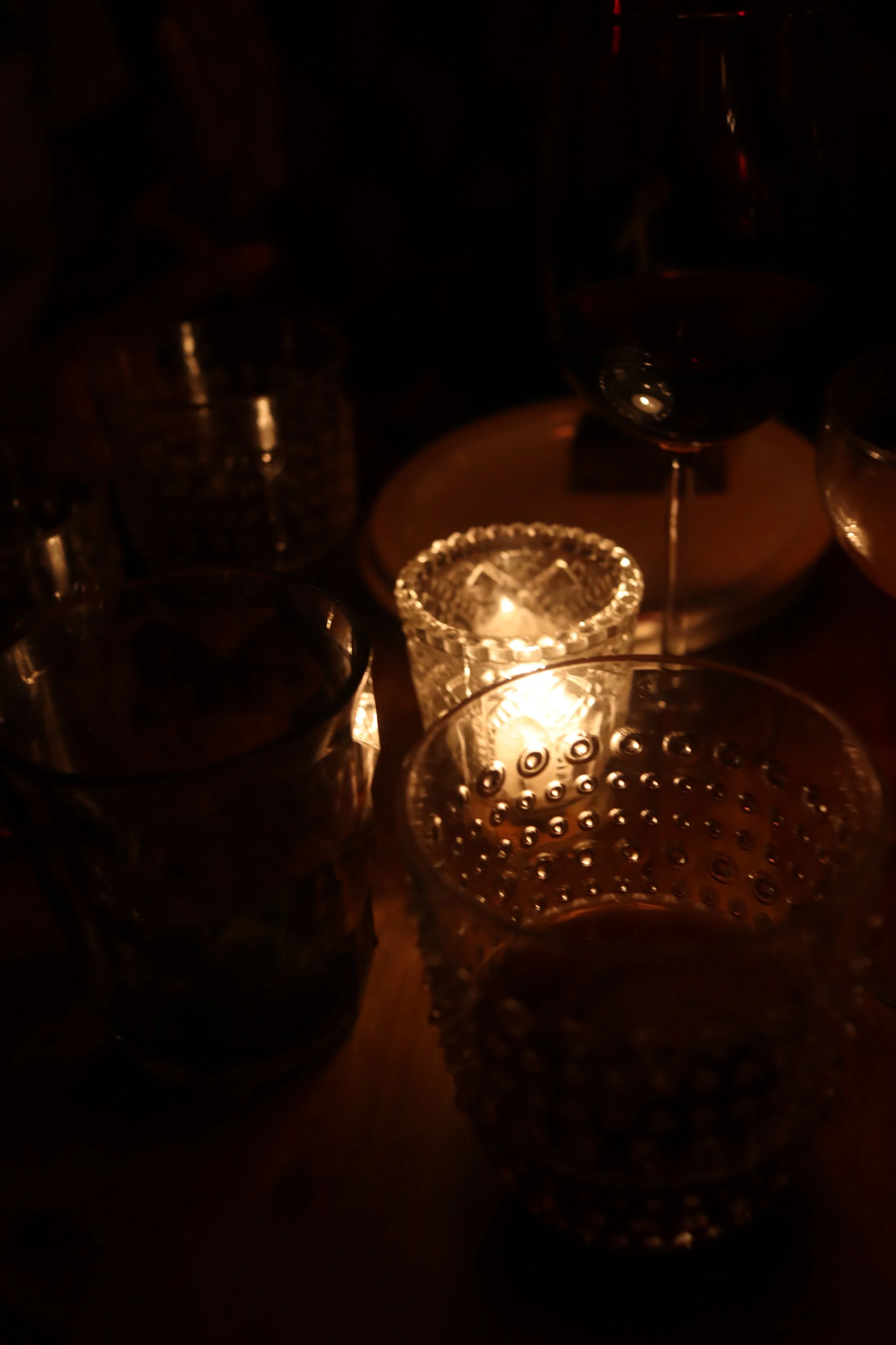 Dimly lit scene with several drinking glasses, and a small lit candle in a textured glass holder on a table.