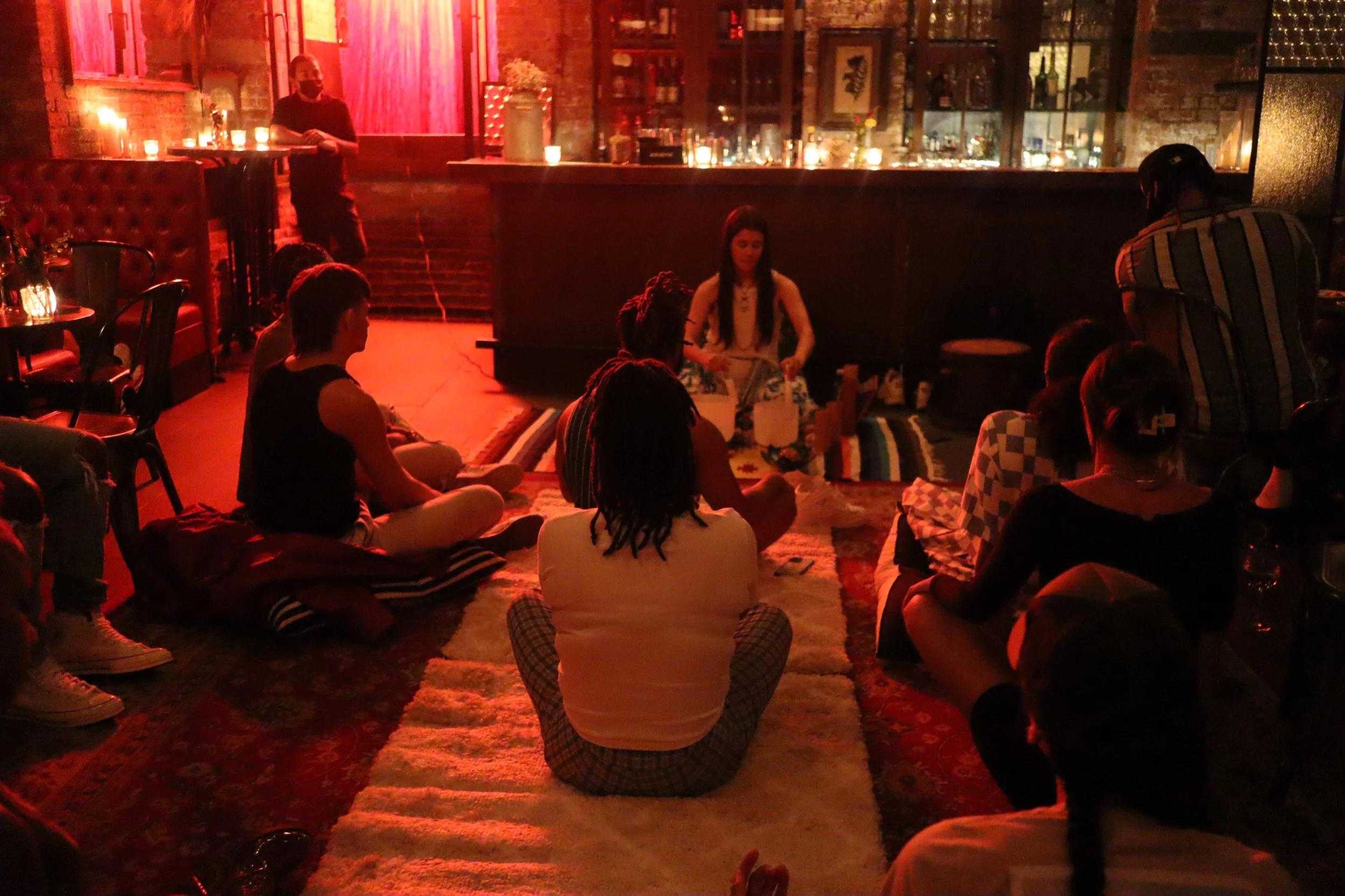 A group of people seated on a rug in a dimly lit indoor space, listening to a woman who is sitting on a blanket, possibly hosting a discussion or storytelling session. The background features a bar and warm lighting.
