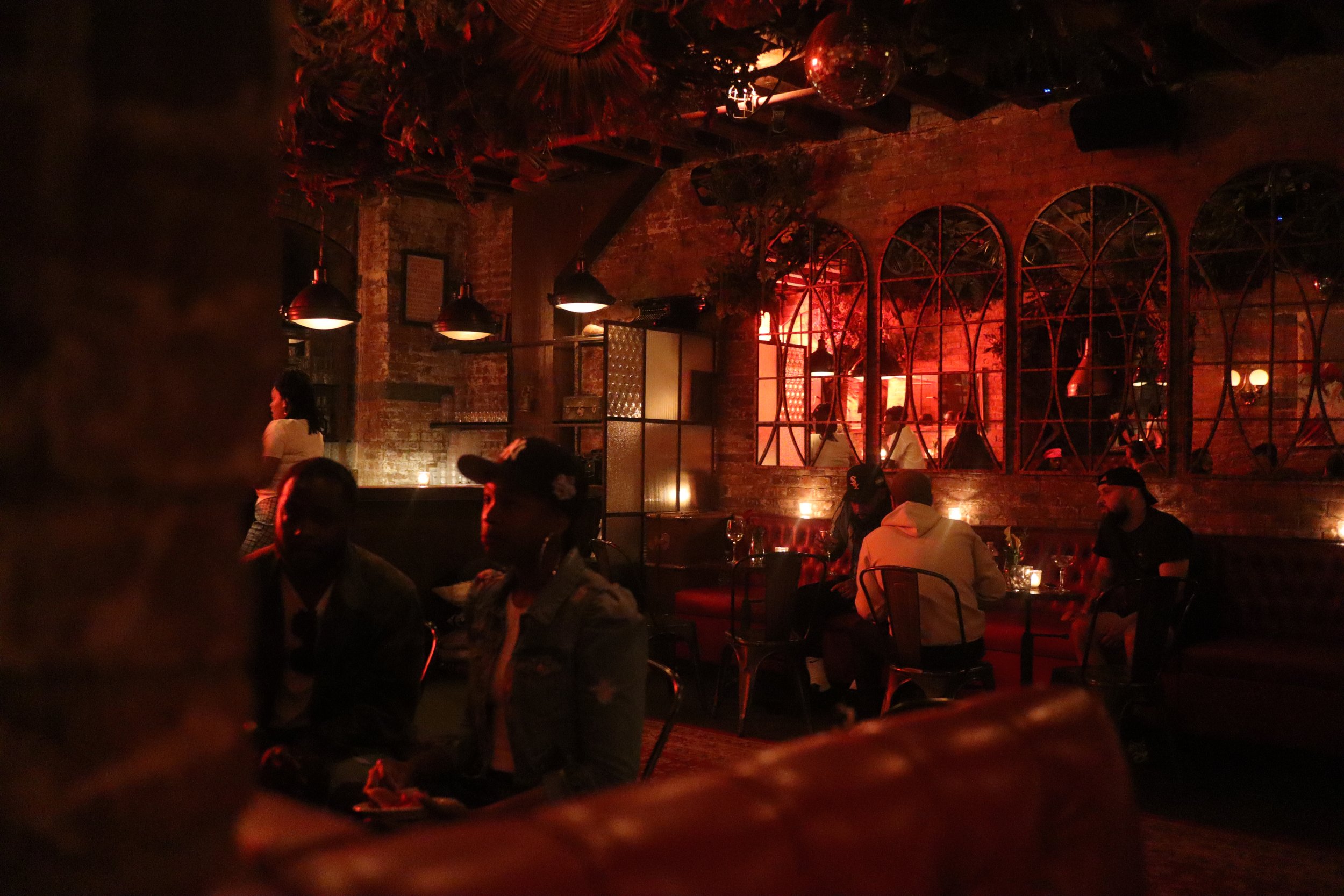 Dimly lit restaurant with exposed brick walls and red lighting. Several patrons are seated at tables, and there is a woman standing near the counter in the background. The decor features arched mirrors and warm, cozy ambiance.