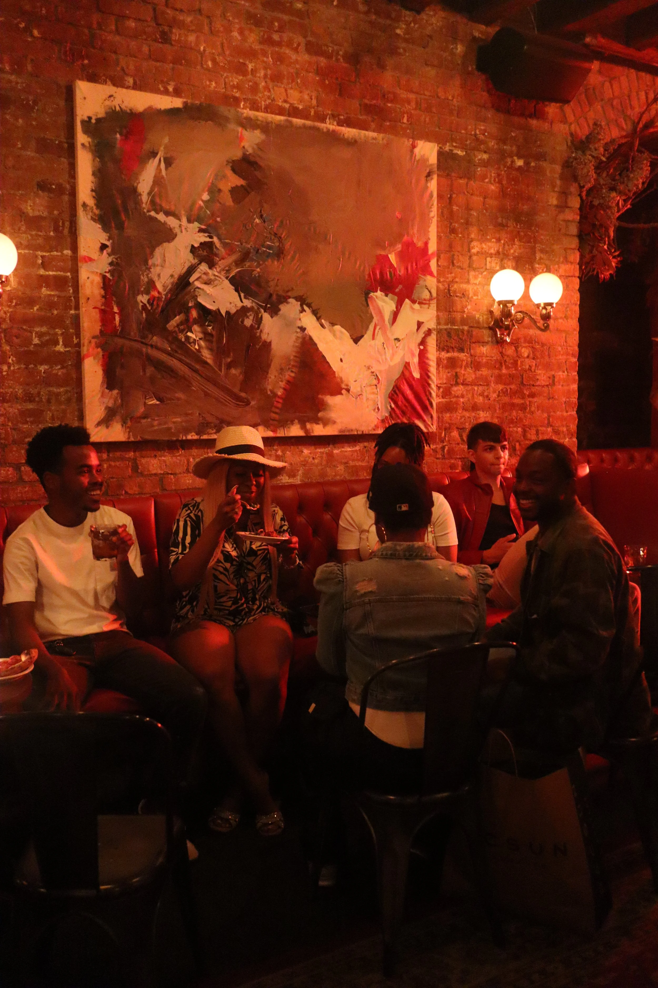 A group of six diverse people sitting on a red leather banquette in a dimly lit restaurant or bar with brick walls, modern art painting, and vintage wall lamps, enjoying drinks and conversation.