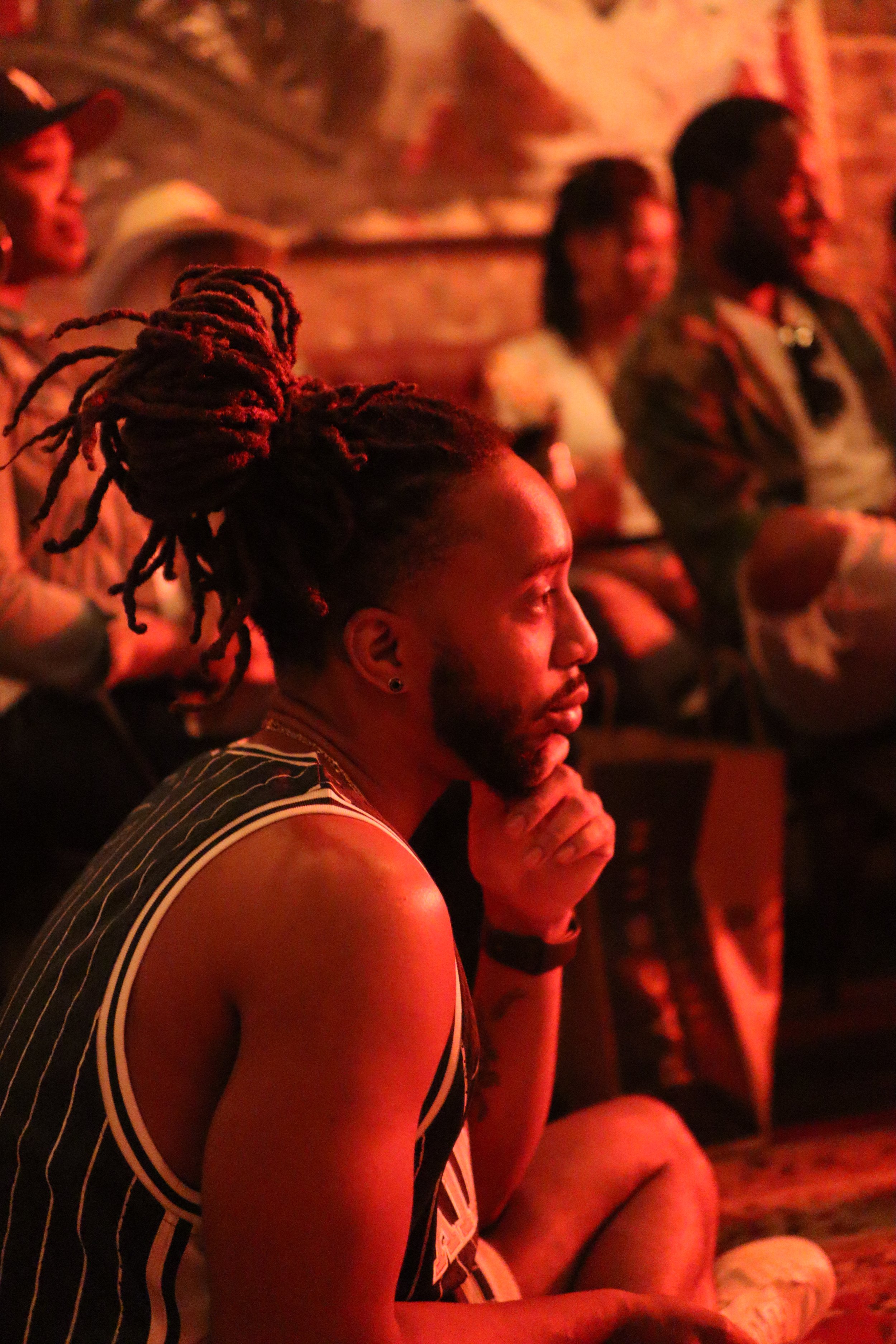 Que sitting with his chin on his hand, attentively watching an event indoors with reddish lighting.