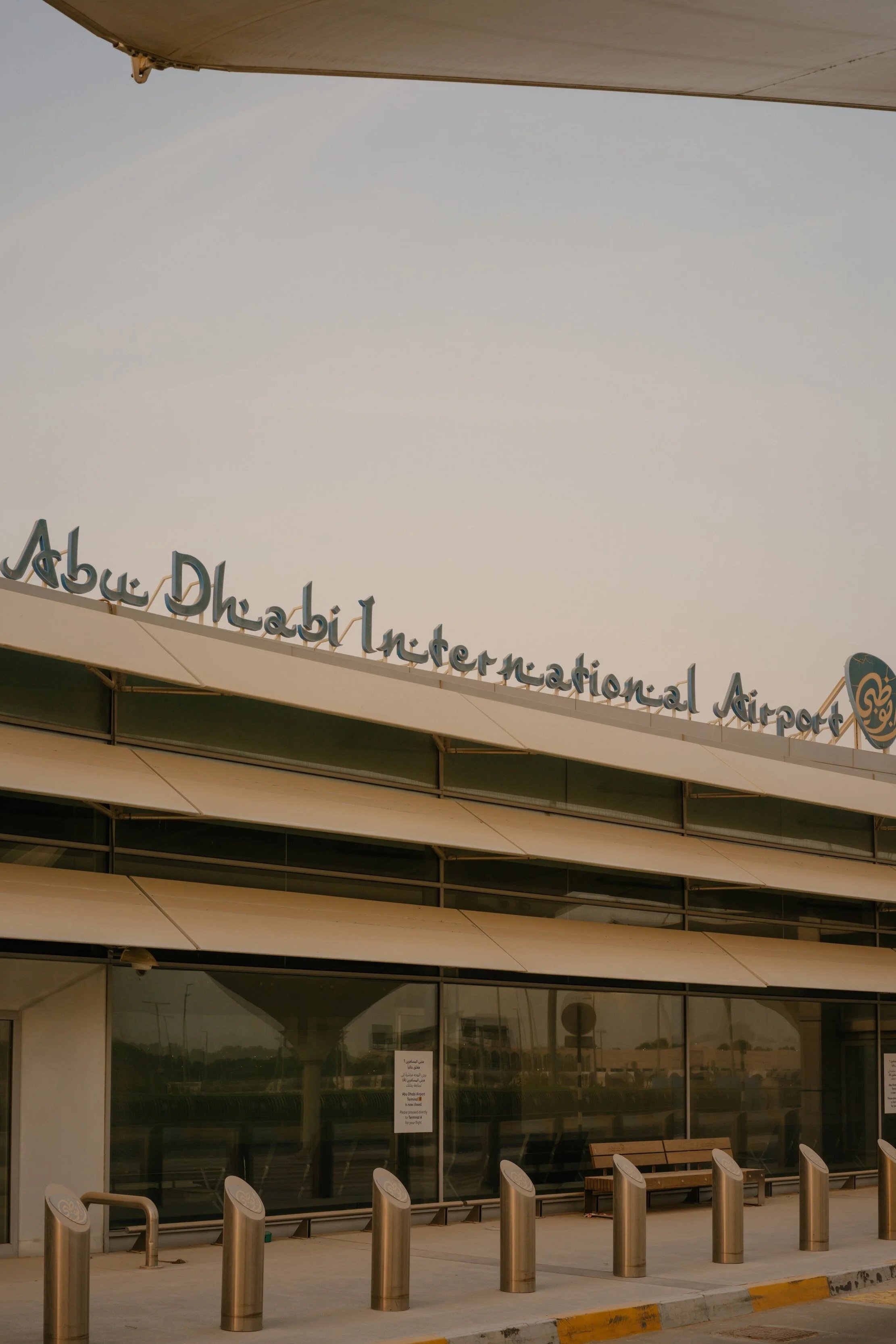 Exterior view of Abu Dhabi International Airport with the airport's name overhead, benches, and metal posts at the entrance.