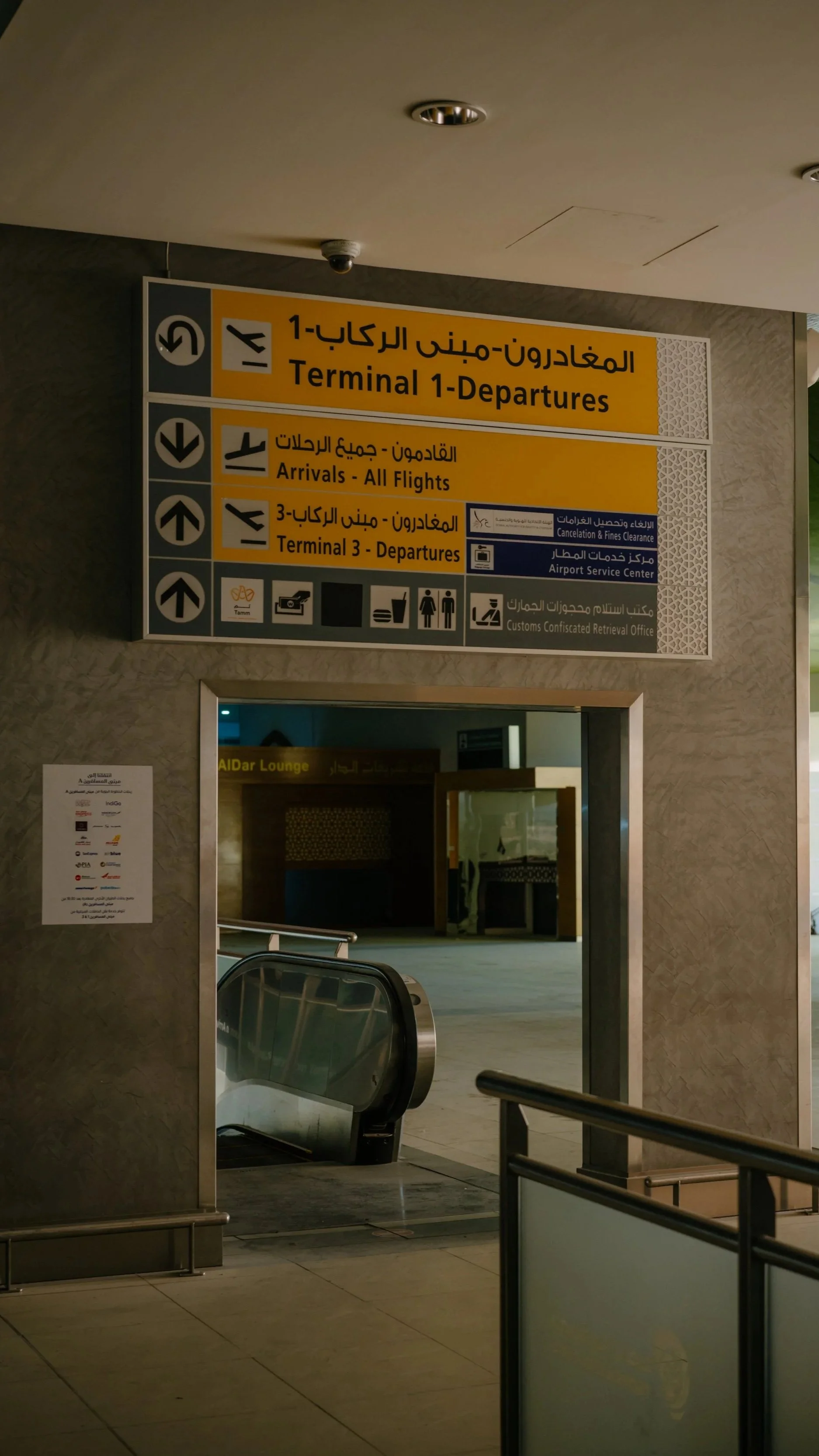 Airport signage indicating directions to Terminal 1 Departures, All Flights arrivals, and Terminal 3 Departures, in English and Arabic, with a view of an escalator and airport waiting area.