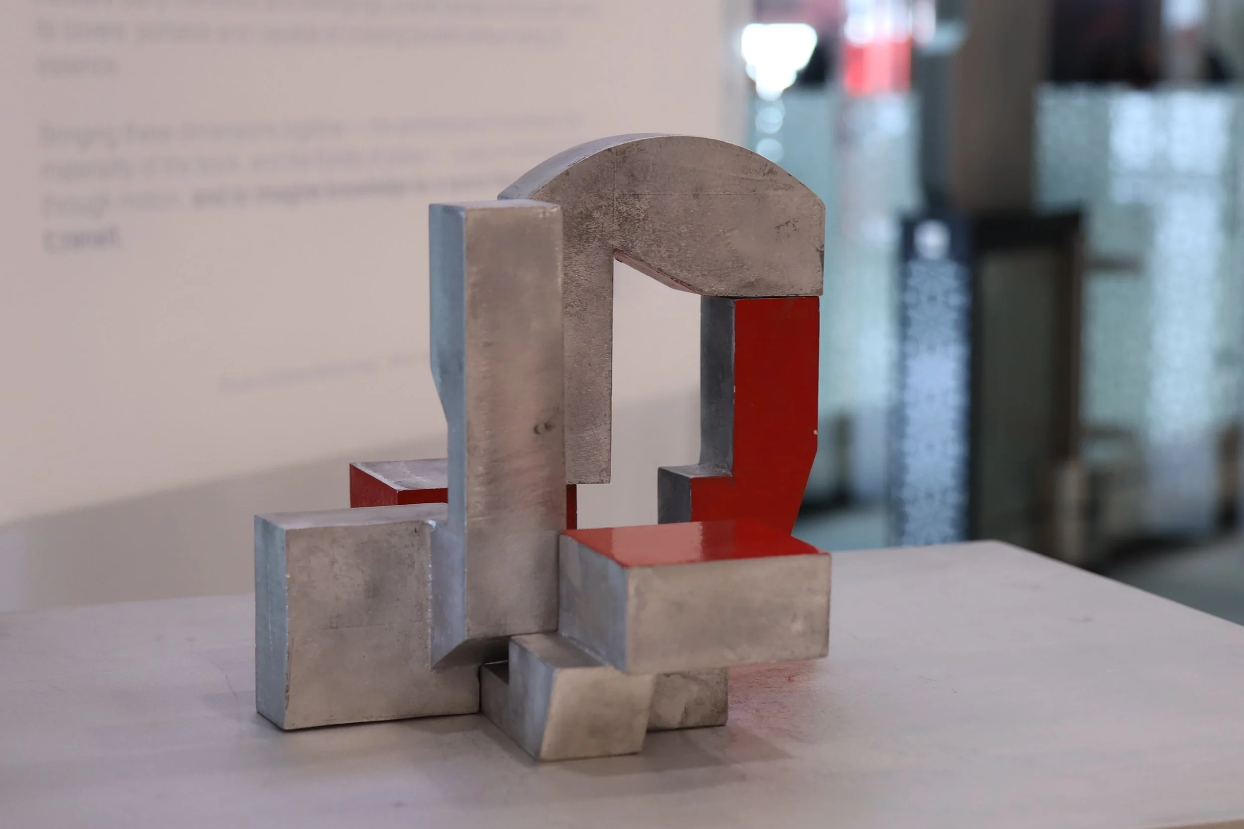Etienne Bastormagi Architecture and Design  - Installation - A Nomadic Library - Abstract metallic sculpture with a geometric intertwined design, some parts painted red, placed on a white surface in an indoor setting.