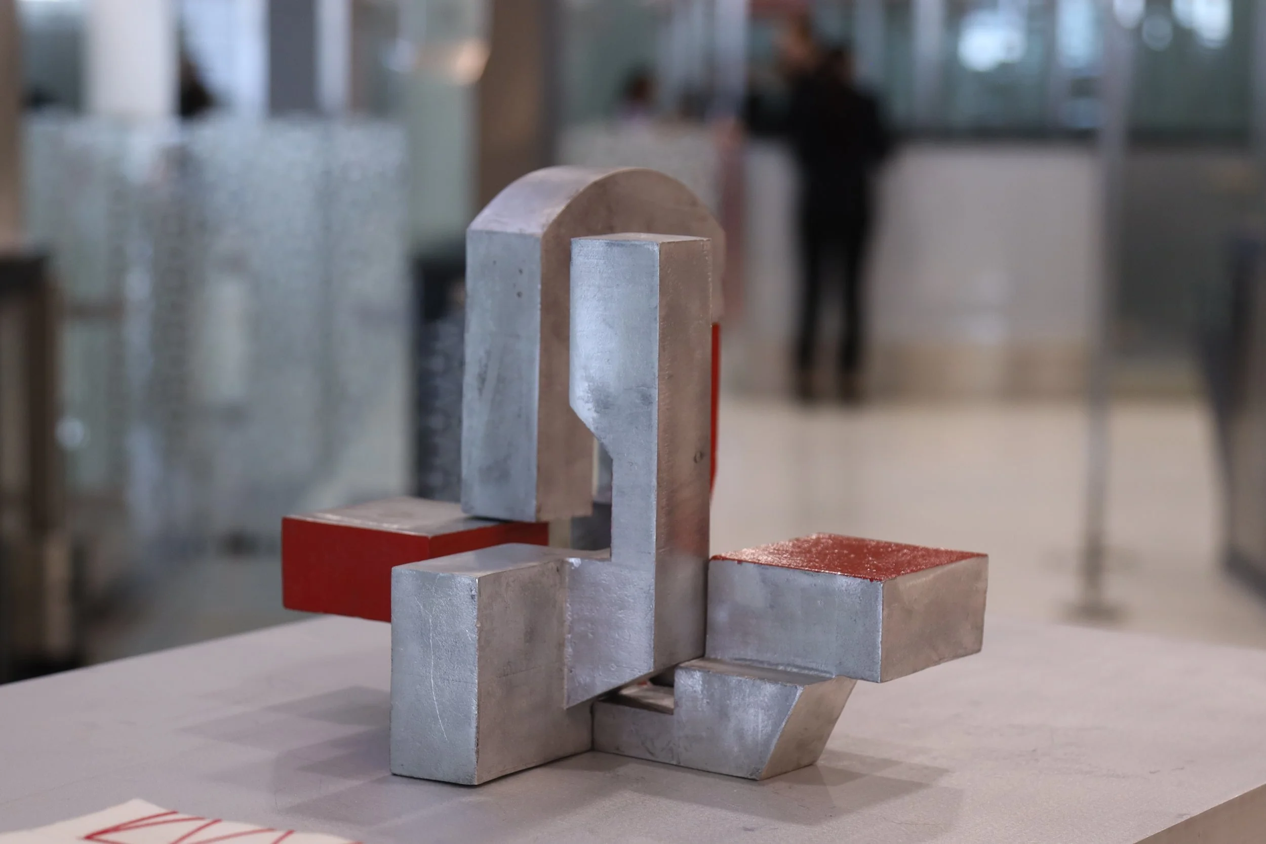 Etienne Bastormagi Architecture and Design  - Installation - A Nomadic Library - Abstract modern sculpture made of metallic blocks with some painted red, placed on a table in an indoor setting with people in the background.
