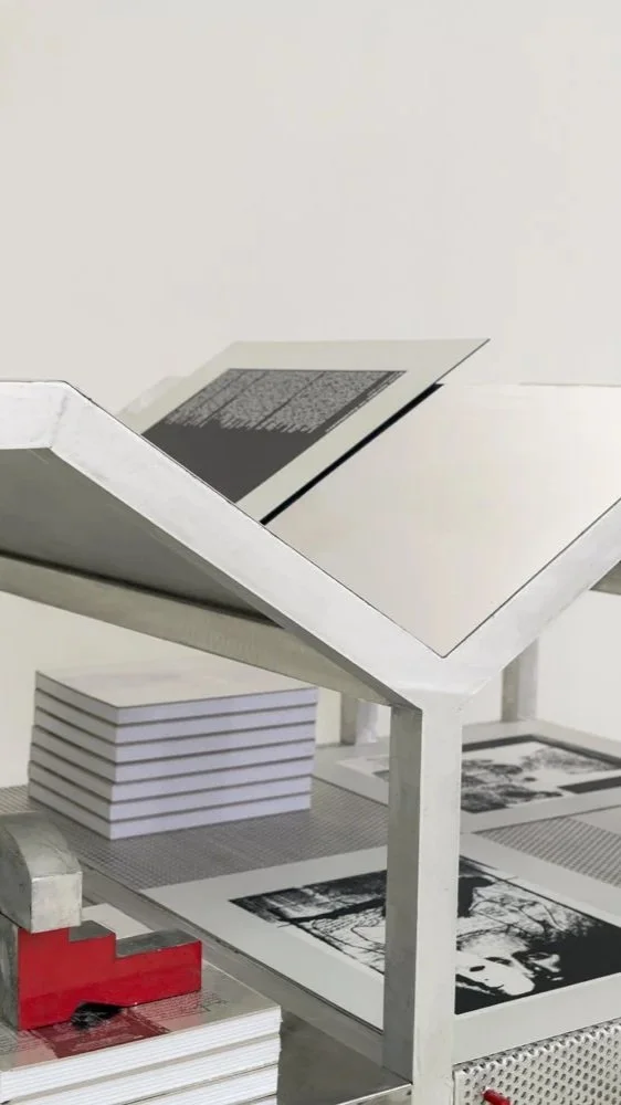 Etienne Bastormagi Architecture and Design  - Installation - A Nomadic Library - Open book or printing press with black and white photographs, stacks of white paper, and a red object on a gray table.