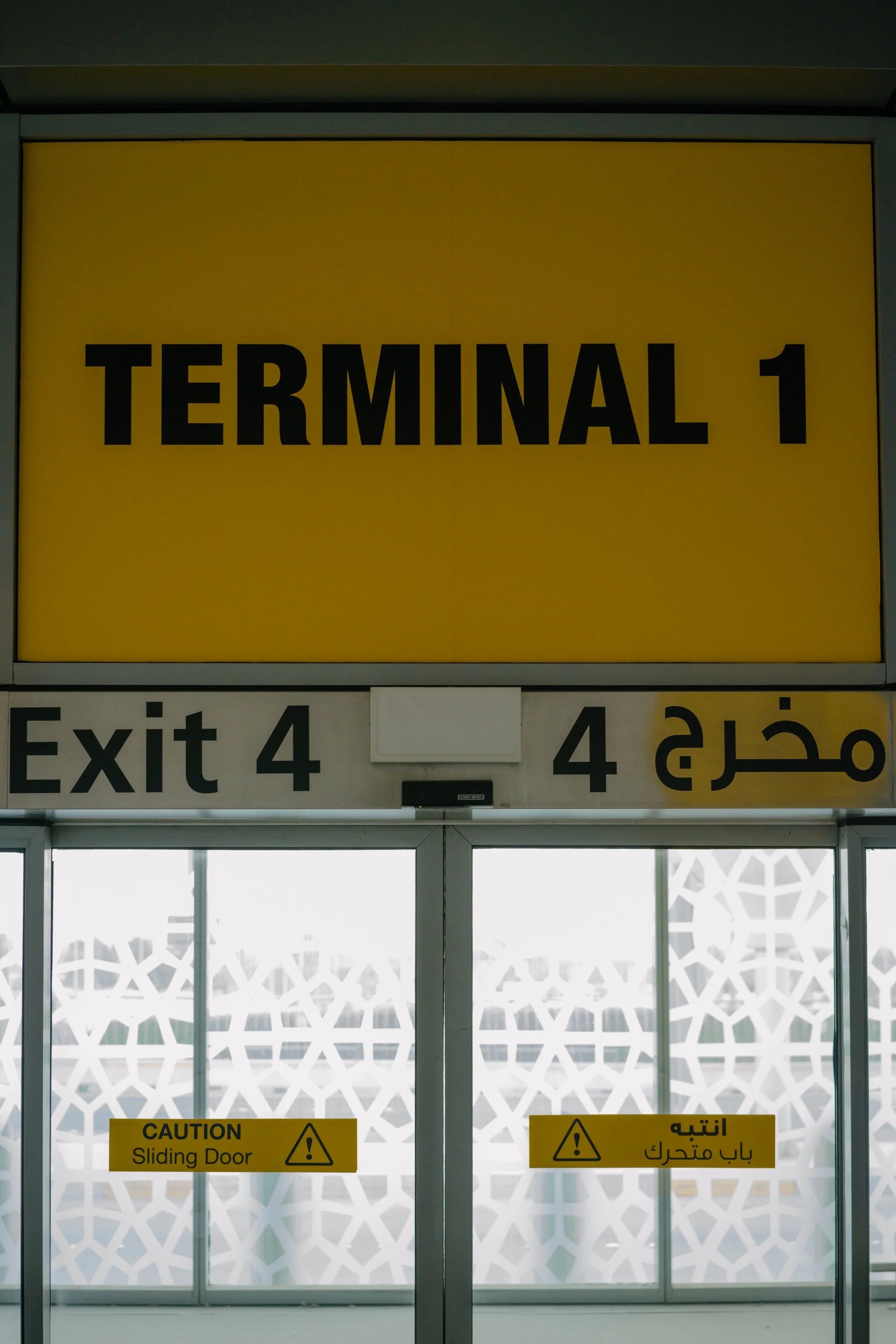 Airport terminal entrance with signs indicating Terminal 1 and Exit 4, including caution signs on sliding doors in English and Arabic.