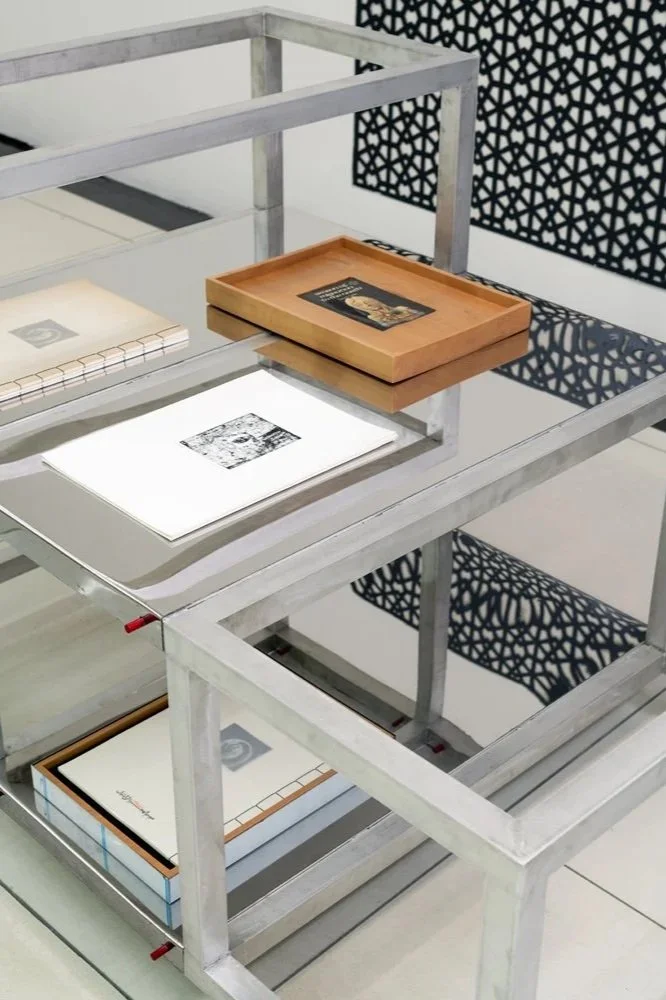 Etienne Bastormagi Architecture and Design  - Installation - A Nomadic Library - A glass-top metal table with two shelves holding books and notebooks, a wooden tray on top of the table, and a decorative black patterned screen in the background.