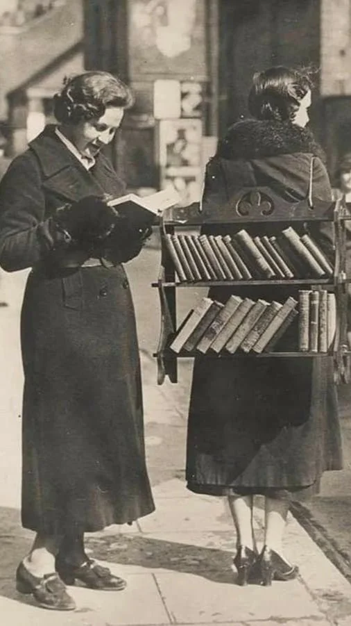 Two women with vintage clothing and shoes stand on a city street, one is reading a book, the other carries a bookshelf filled with books on her back, in a black and white photo.
