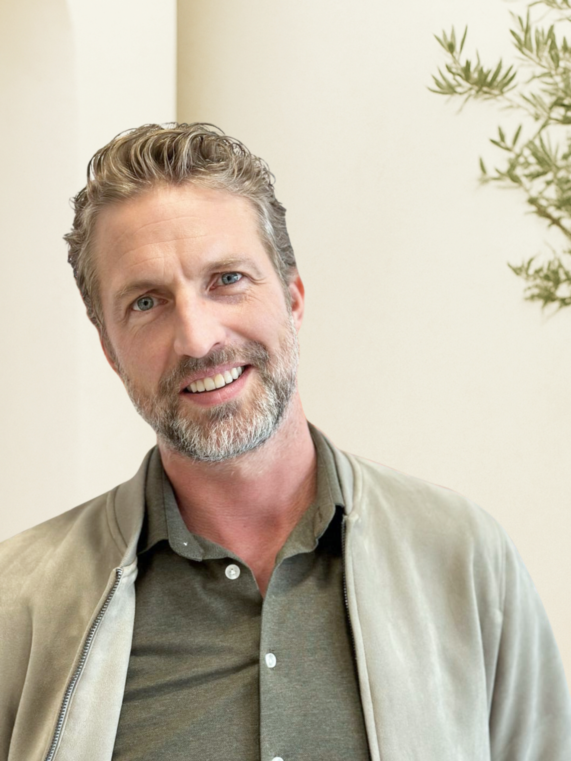 Close-up photo of a smiling middle-aged man with gray hair and beard, wearing a gray button-down shirt and a light gray jacket, standing in front of a light-colored wall with some green foliage visible on the right side.
