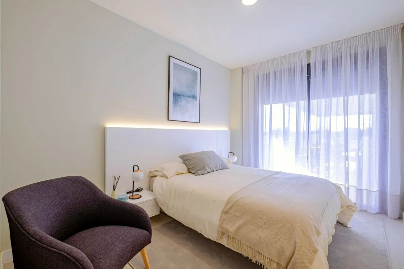 Bedroom with a bed, purple chair, large window with sheer curtains, artwork, and minimalist decor.