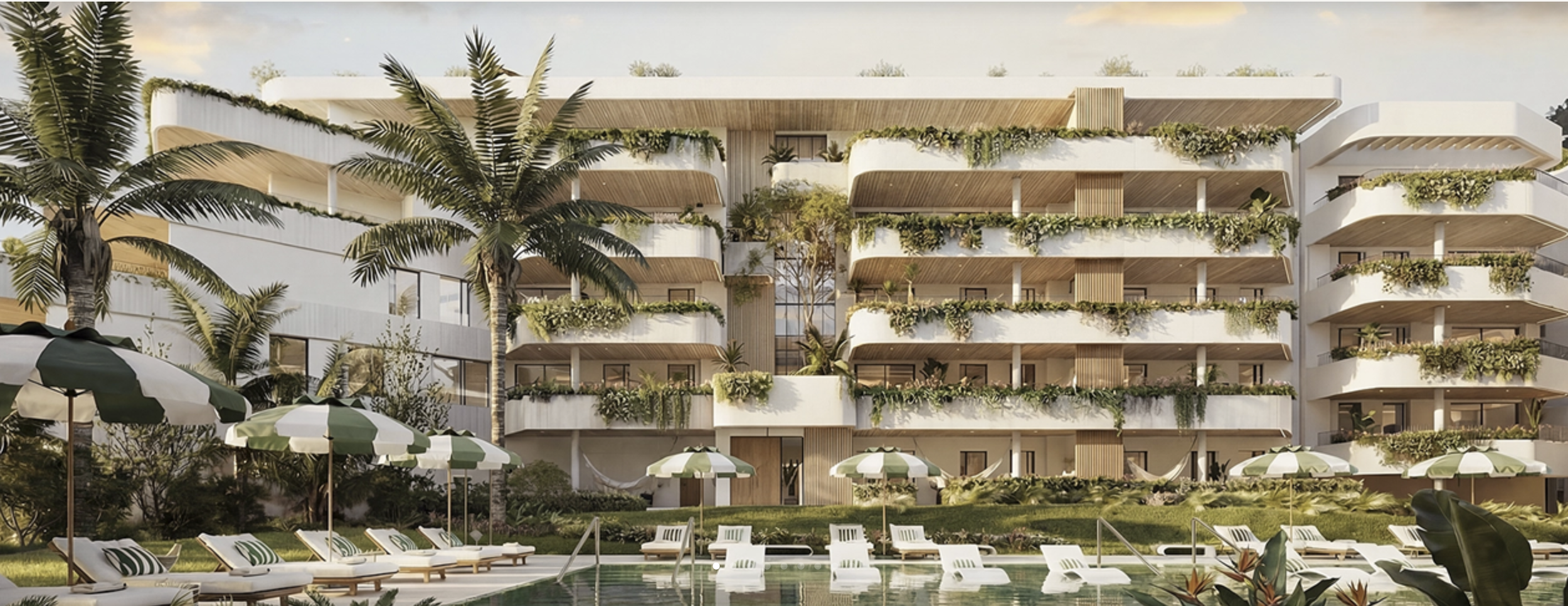 Modern white apartment building with multiple balconies decorated with green plants, surrounded by palm trees and an outdoor pool area with lounge chairs and umbrellas.