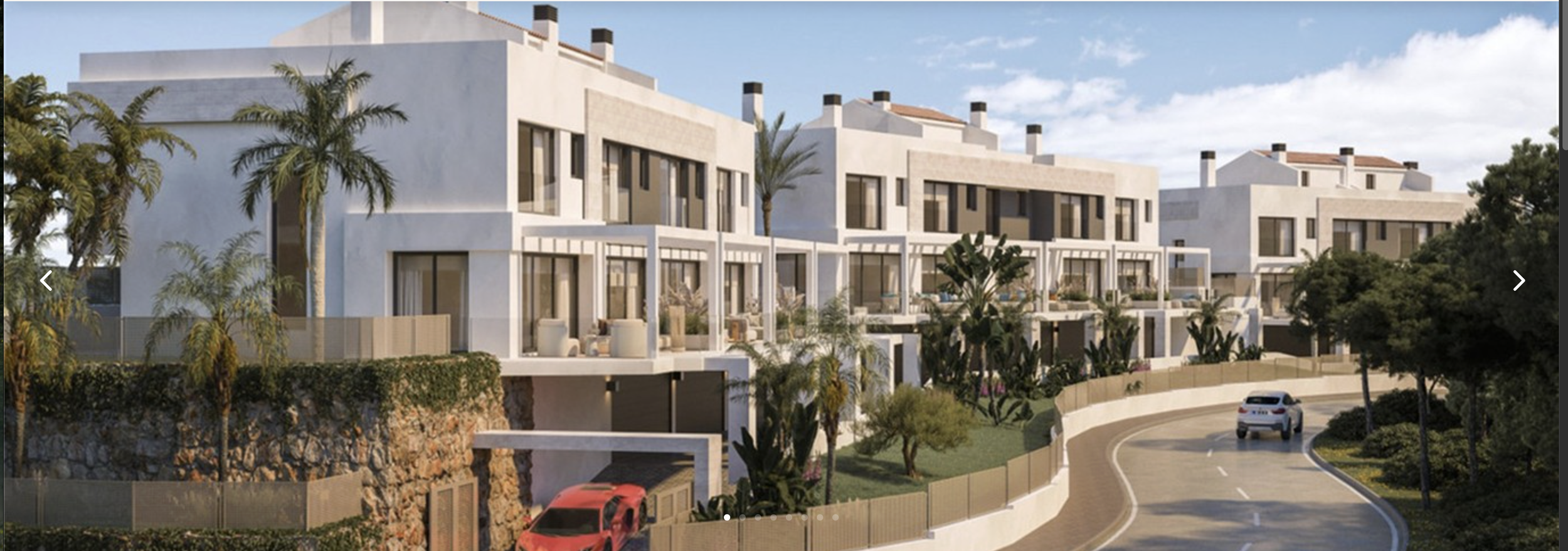 Modern white residential buildings with balconies and large windows, surrounded by trees and a winding road with parked cars.