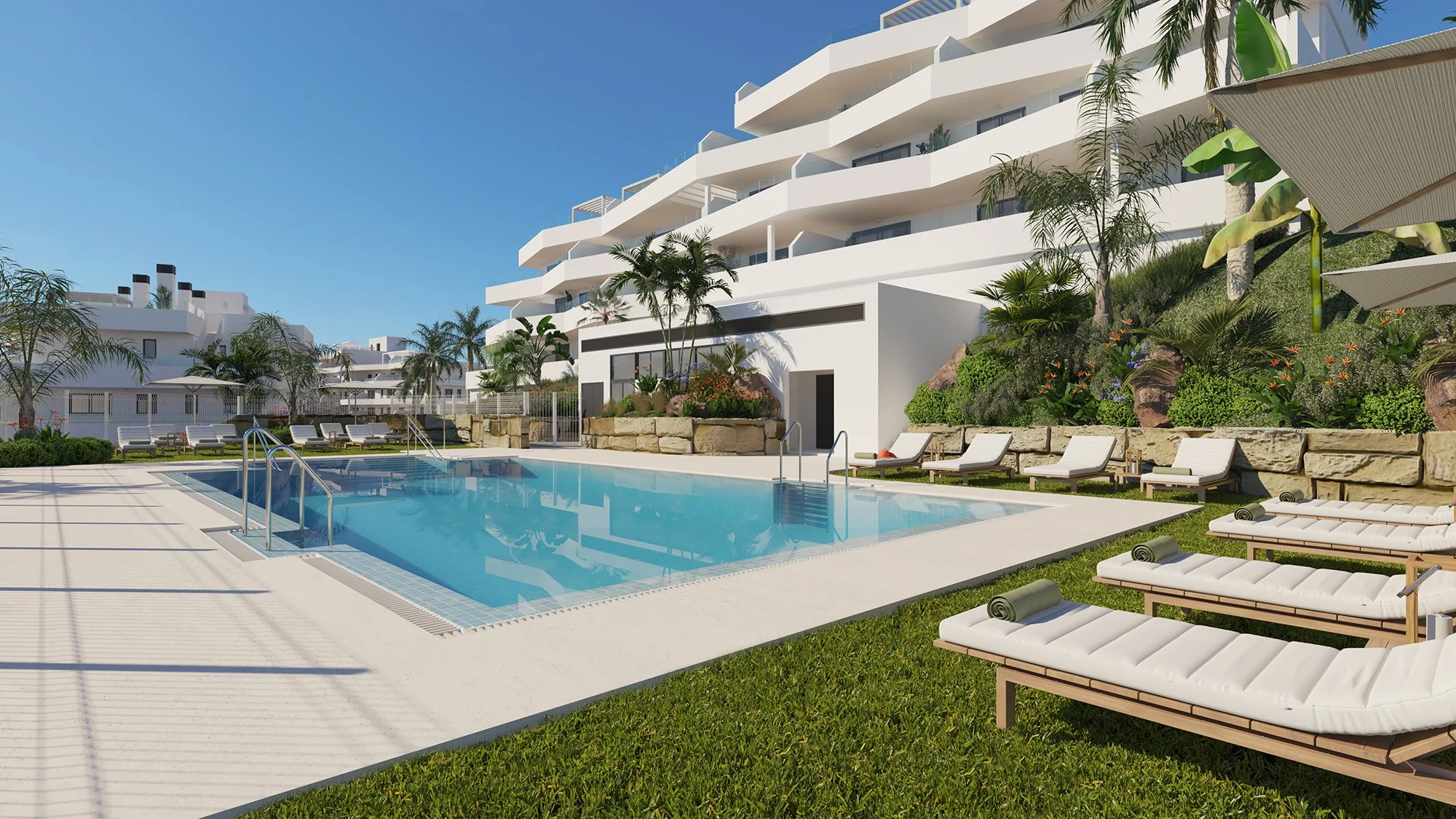 Luxury apartment complex with a swimming pool surrounded by white lounge chairs, palm trees, and modern white buildings under a clear blue sky.