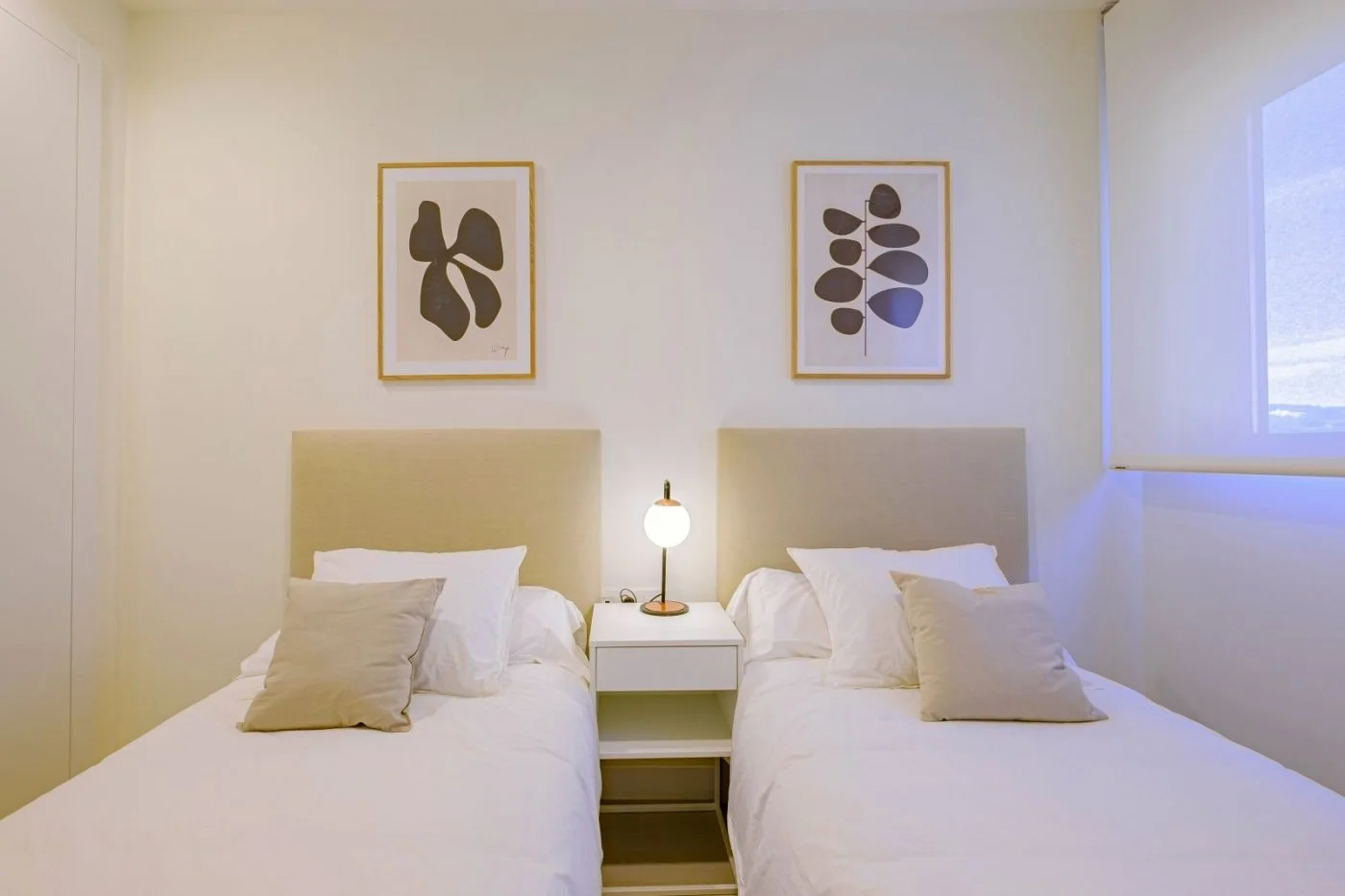 A contemporary bedroom with two beds, each with white bedding and beige pillows, separated by a nightstand with a round table lamp, above which are two abstract art pieces on the wall and a window with a roller blind.
