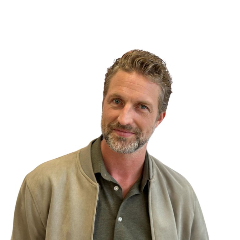 A man with short, wavy hair and a beard, wearing a gray collared shirt and a beige jacket, looking at the camera with a slight smile.