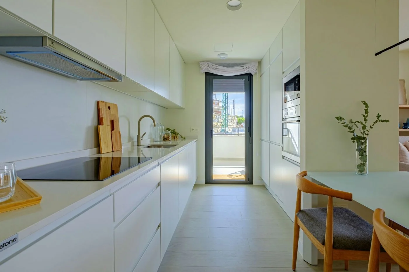 Modern kitchen with white cabinets, a black induction stove, a wooden cutting board, and a sliding glass door leading to a balcony with a view of the outdoors.