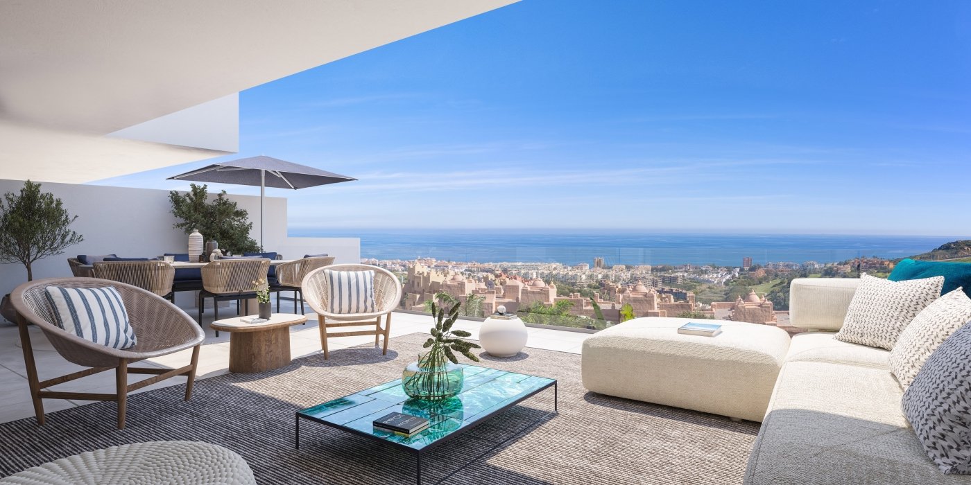 Luxury outdoor terrace overlooking a city and ocean view, furnished with a white sectional sofa, patterned cushions, wicker chairs with striped pillows, a glass-topped coffee table with a plant, and an umbrella providing shade.
