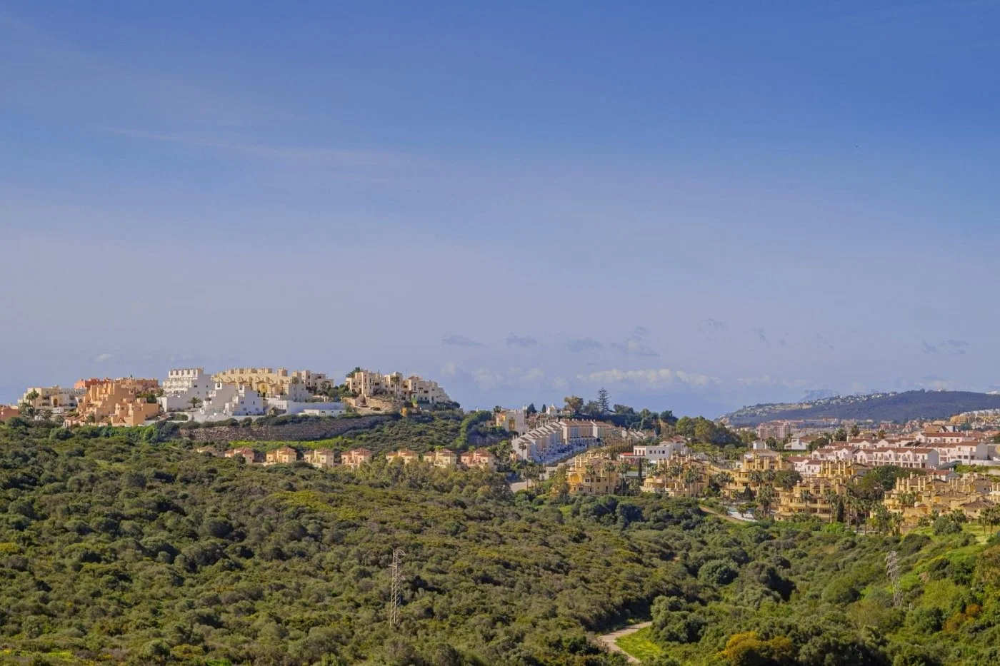 A hilly landscape with residential buildings atop the hills and lush green vegetation covering the slopes, under a clear blue sky.