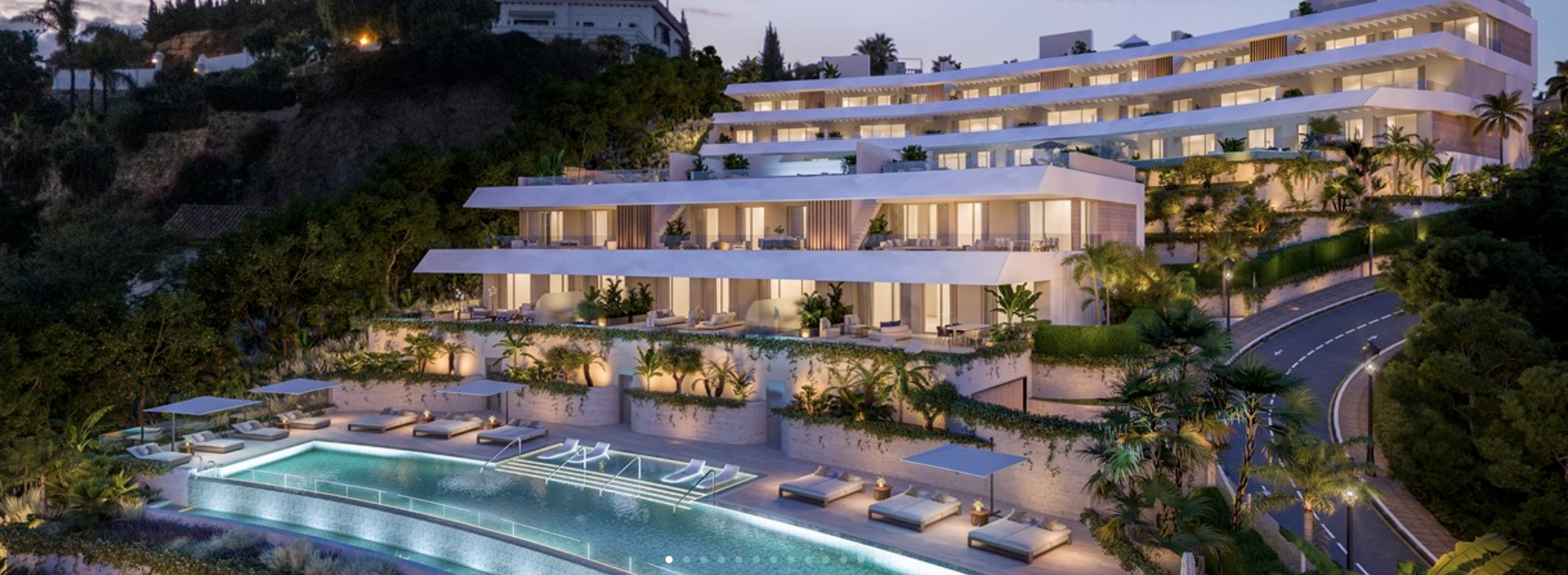 Night view of a modern multi-story hillside hotel with balconies, illuminated exterior lighting, palm trees, and a swimming pool with lounge chairs and umbrellas.