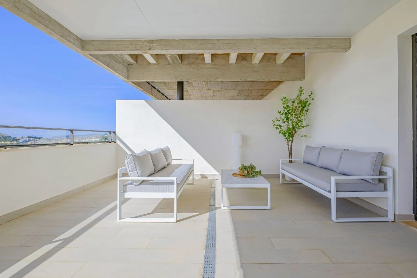 Balcony with white outdoor furniture, including two sofas and a coffee table, a potted plant, and a view of a city skyline in the distance under a blue sky.