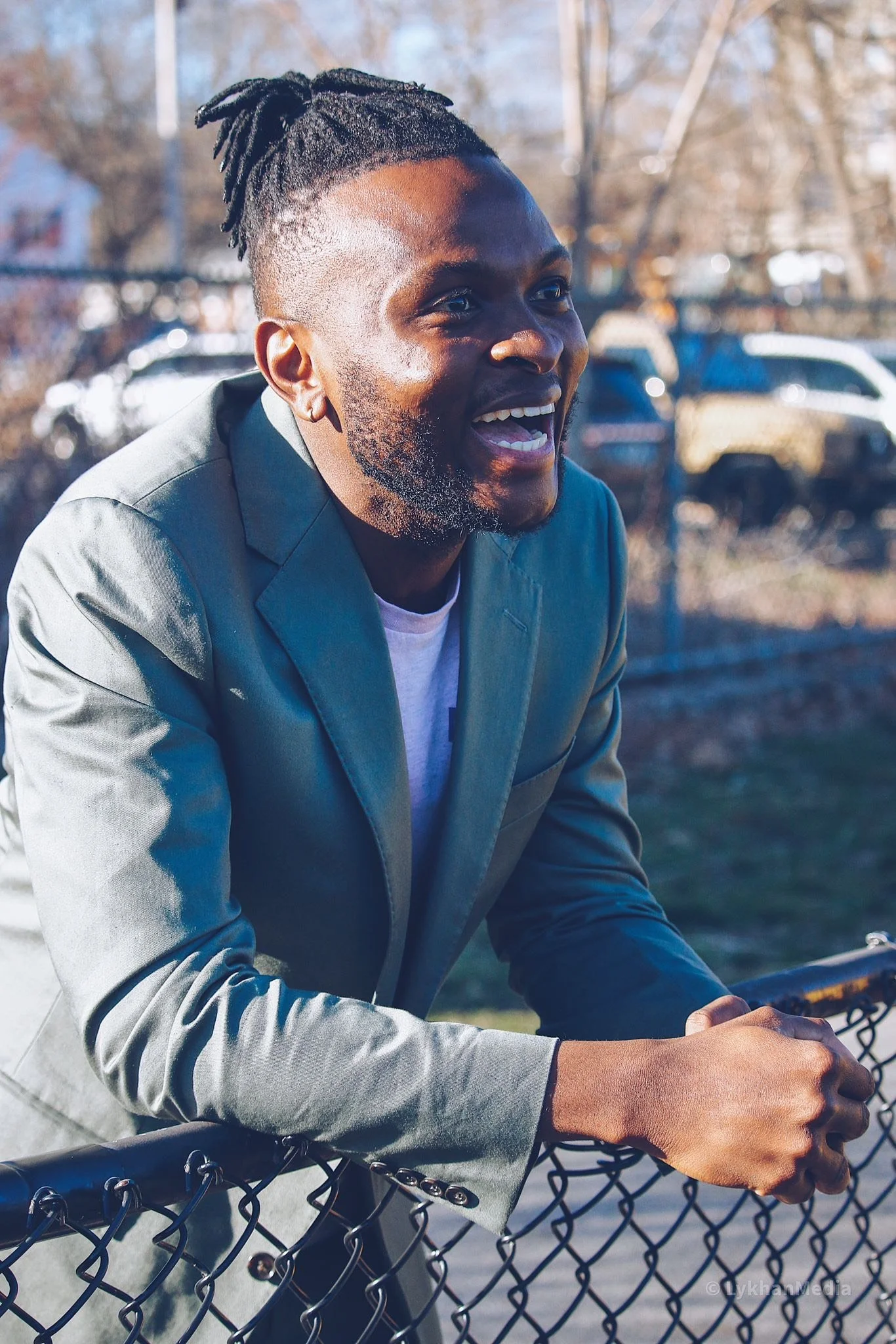 A man with dreadlocks smiling outside in front of a chain-link fence, with cars and trees in the background.