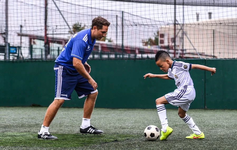 A young boy in a white soccer uniform practicing dribbling a soccer ball on the field while an adult coach in a blue shirt and shorts offers guidance during training.