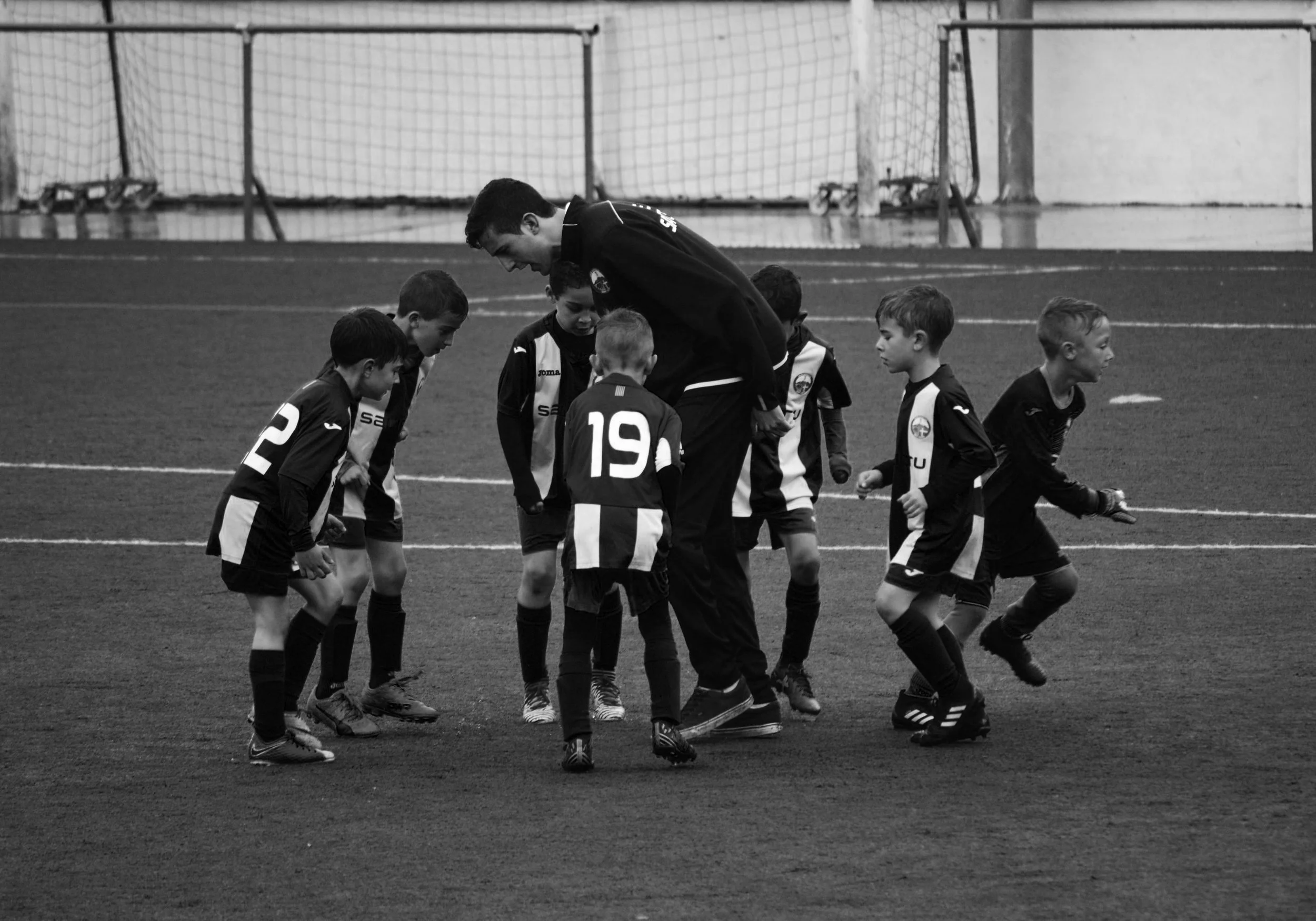 Black and white photo of young boys in soccer uniforms walking on a field with a coach bent over talking to them.