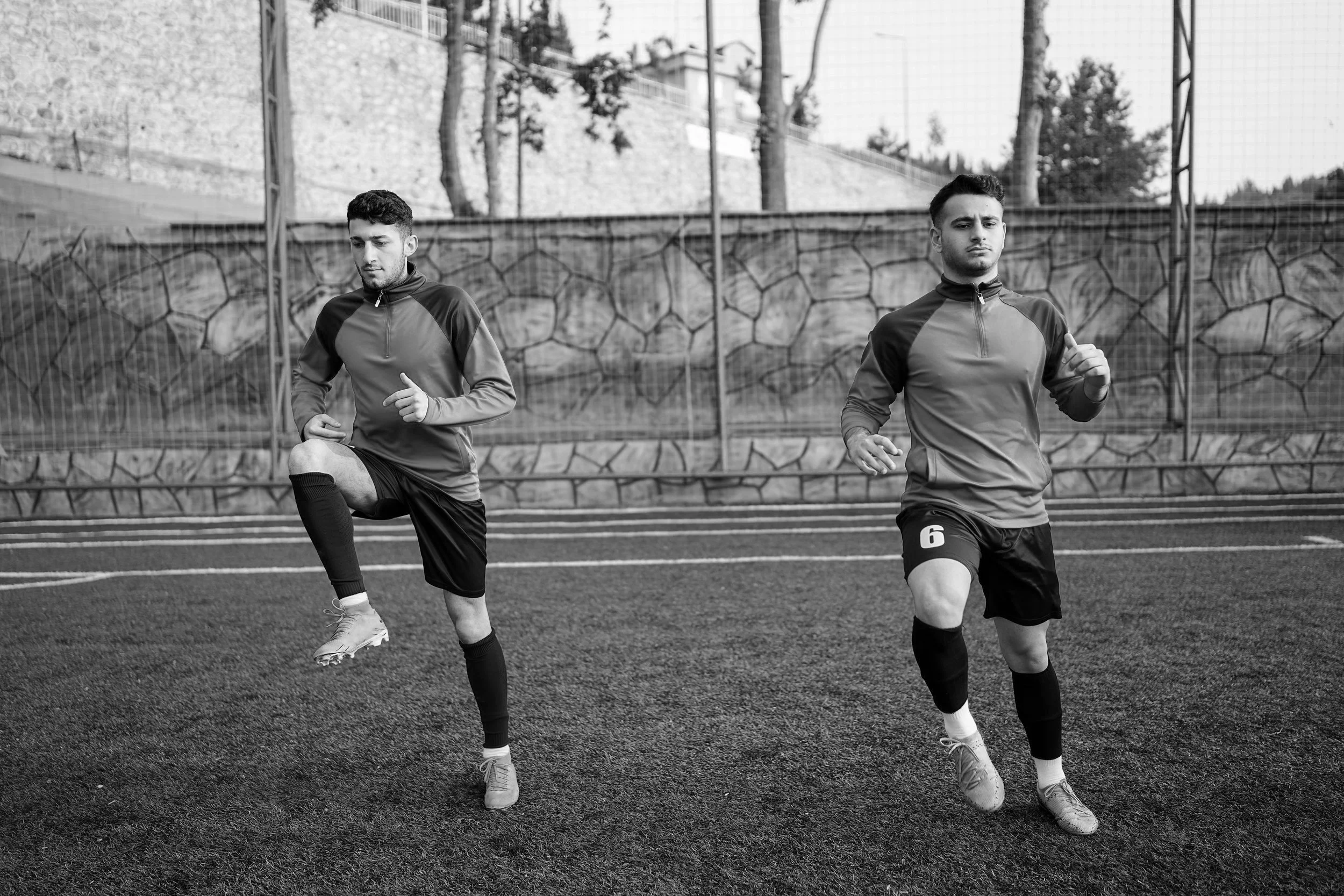 Two male soccer players warming up on an outdoor field, stretching their legs before a game, with a stone wall and trees in the background.
