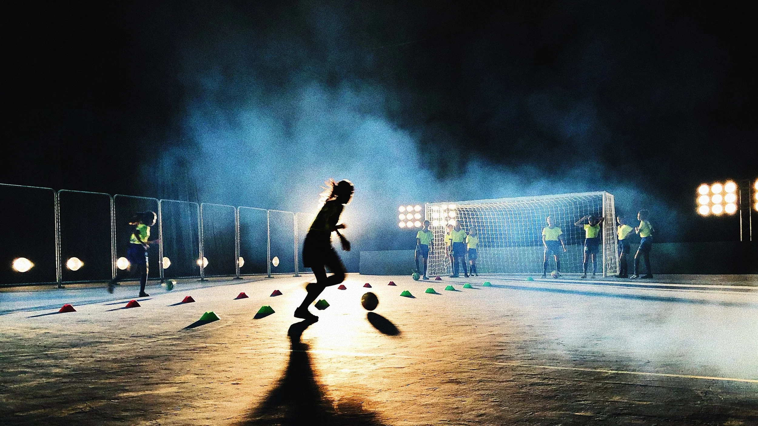 Young women practicing soccer on an illuminated court at night, with cones set up for drills and a goal in the background.