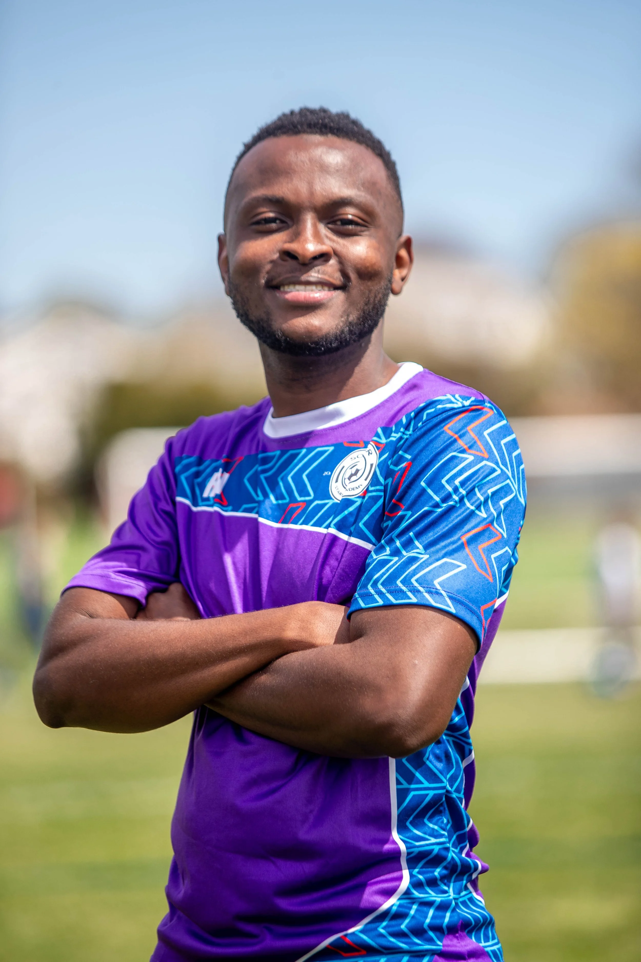 Smiling male athlete wearing a purple and blue sports jersey with crossed arms outdoors on a sunny day.