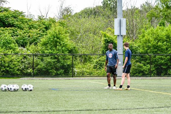 Two boys standing on a soccer field near a fence, with soccer balls on the ground nearby, surrounded by trees.
