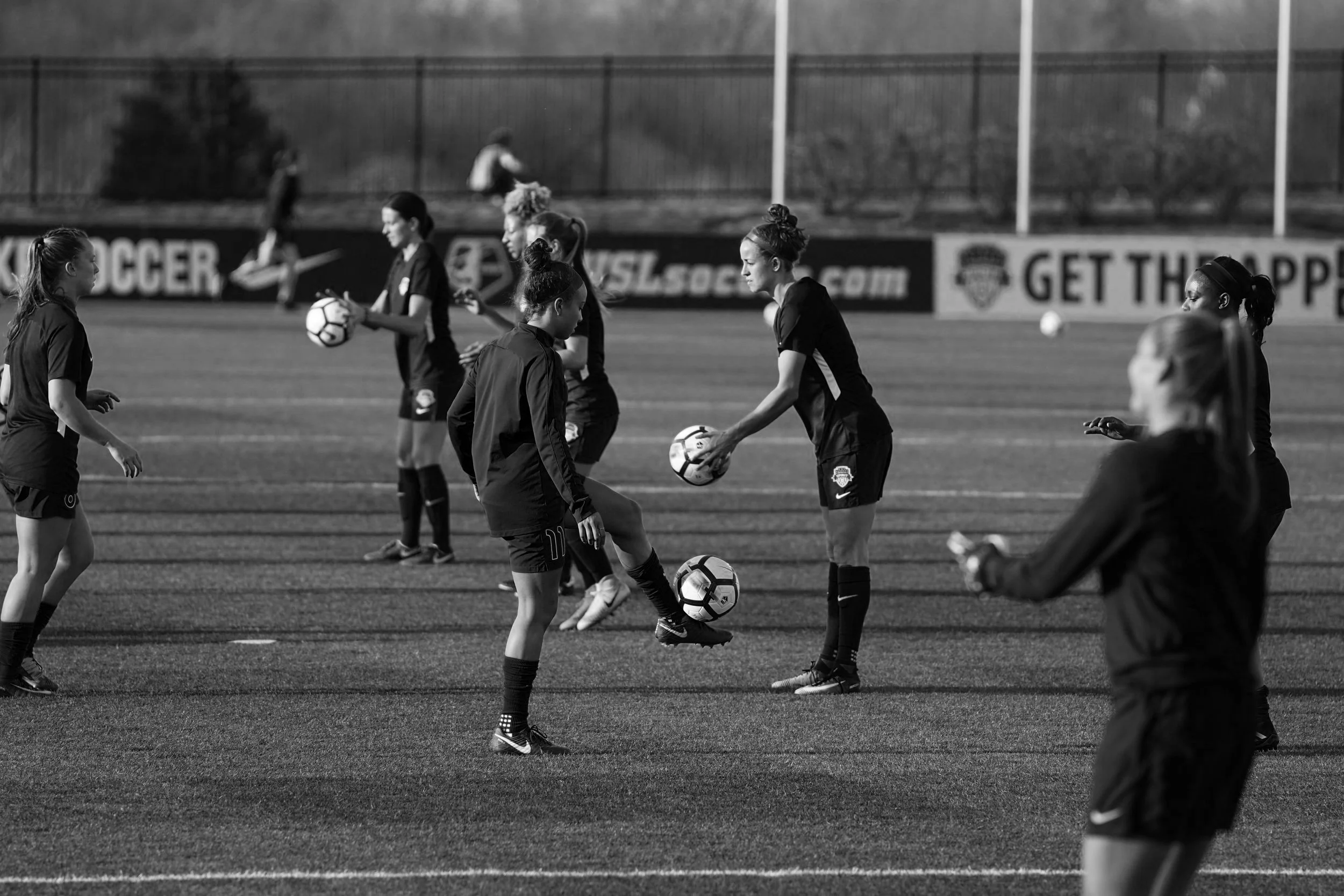 Group of female soccer players on the field during practice or warm-up, some holding soccer balls, in a mostly empty stadium with advertising boards in the background.