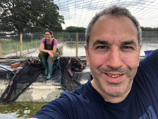 A man takes a selfie in front of a woman sitting on a ledge at a fish hatchery or outdoor fish pond, with fishing nets and bags around them.