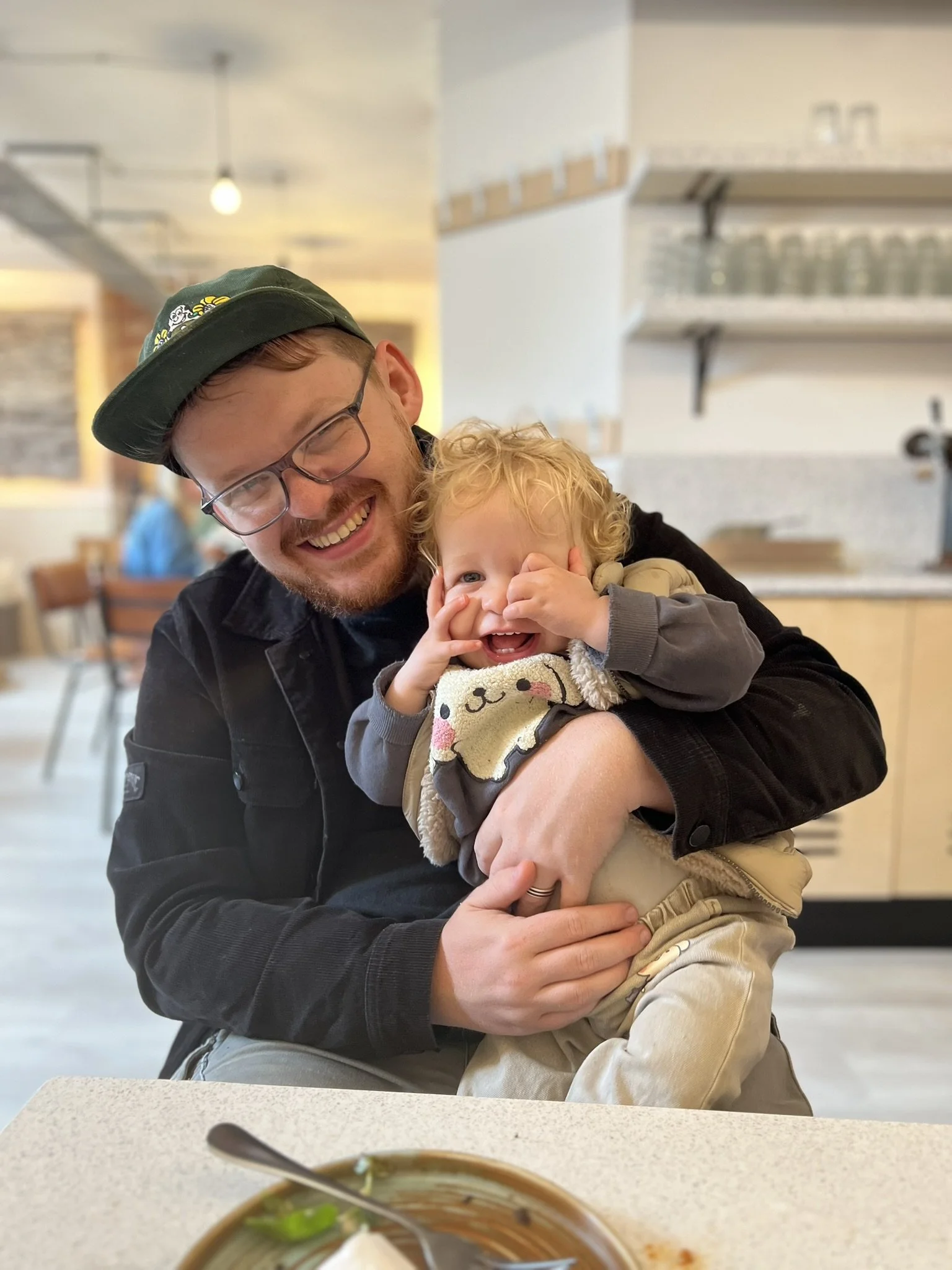 A man with glasses and a hat holding a smiling blonde child in a restaurant.