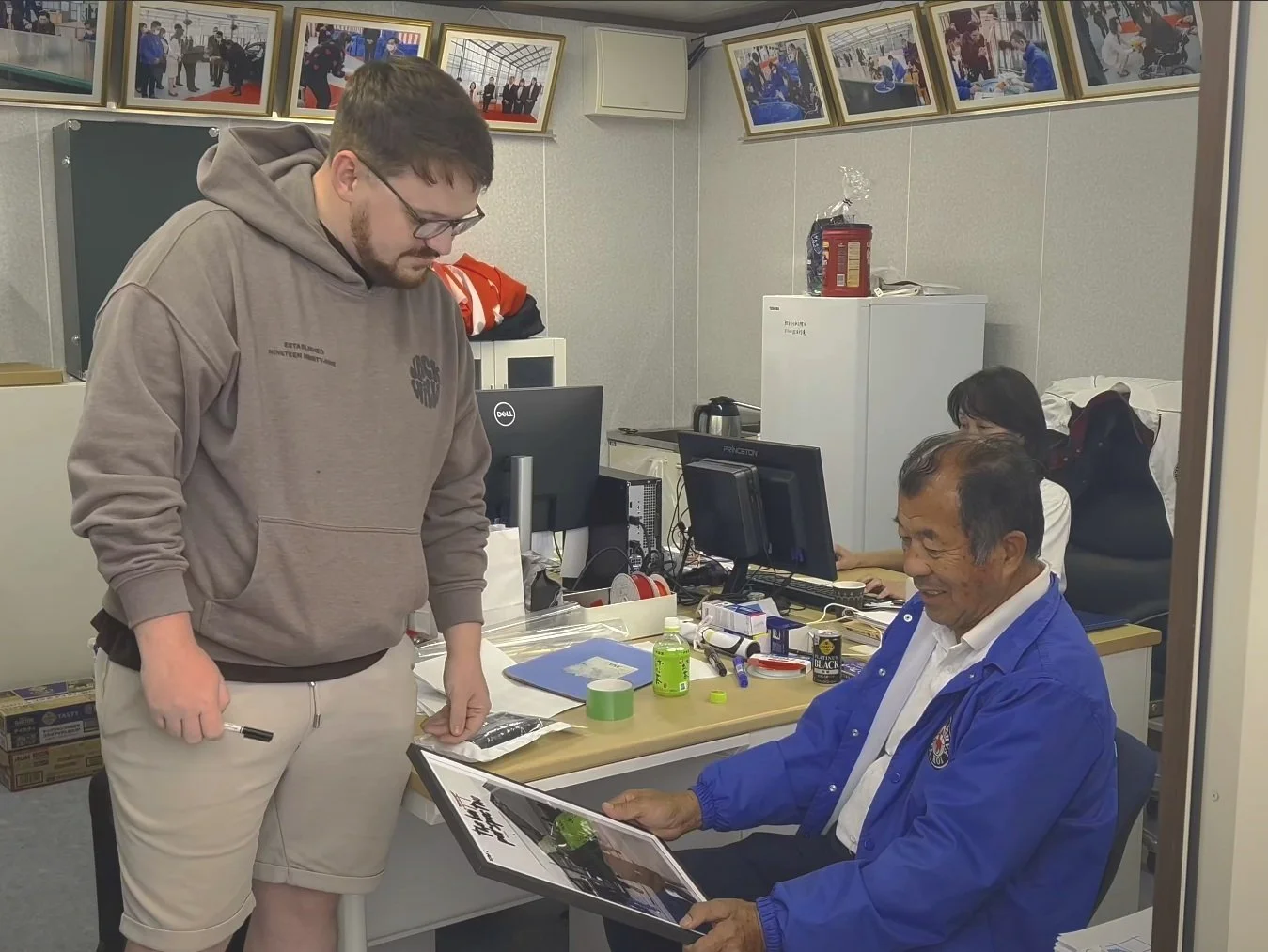 A young man in a brown hoodie and beige shorts looking at an elderly man in a blue jacket who is holding a photo album, in an office setting with two women working at computers in the background and framed photos on the wall.