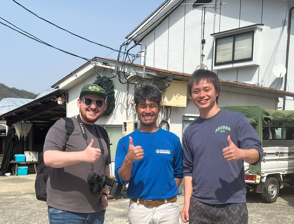 Three men smiling and giving thumbs up outdoors in front of a house and vehicles.