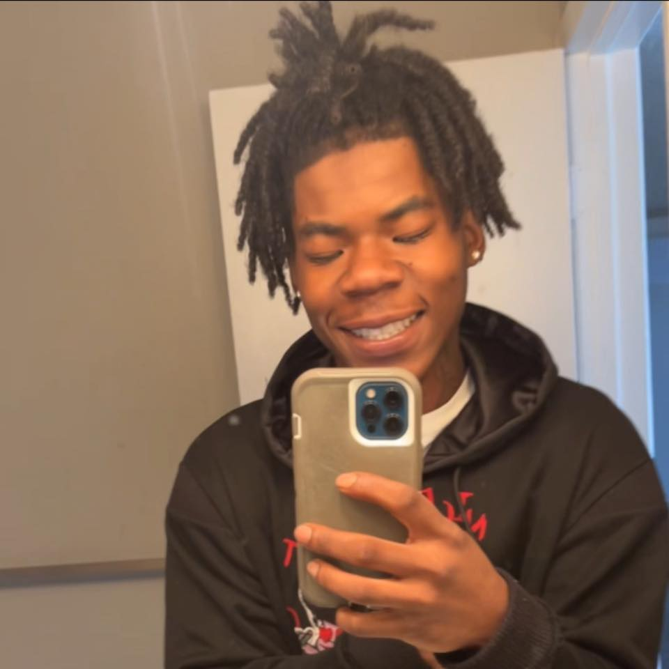 A young man with dreadlocks taking a mirror selfie, smiling, wearing a black hoodie.