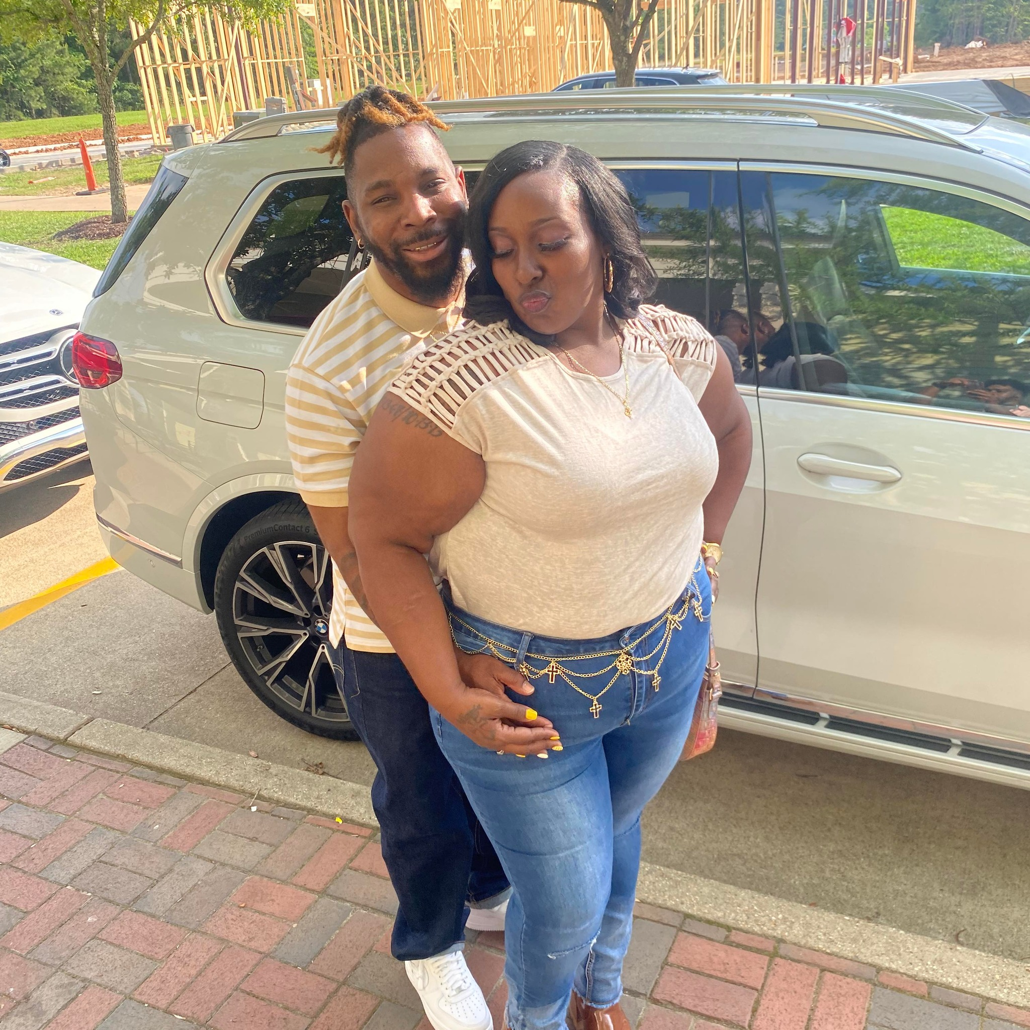 A man and woman pose together outside near a silver SUV, with construction and trees in the background, the woman making a pout face and the man smiling behind her.