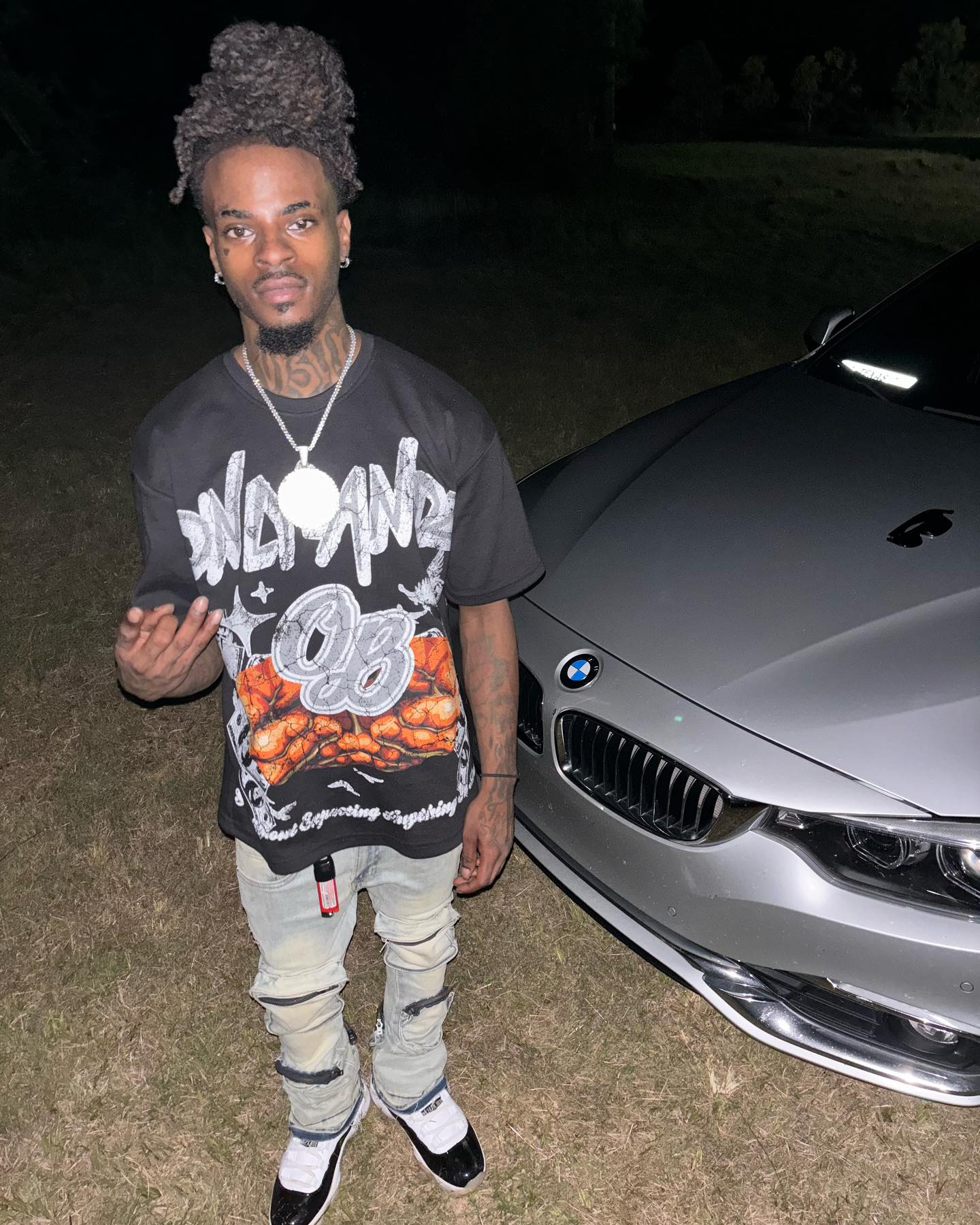 A man with dreadlocks, tattoos, and jewelry standing next to a silver BMW car at night.