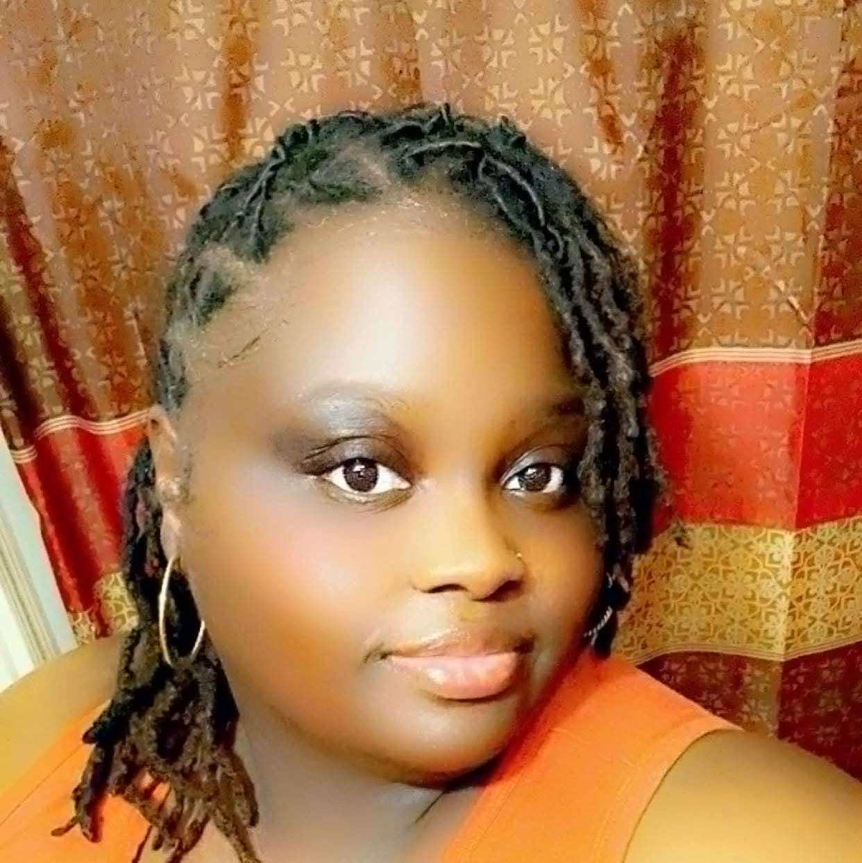 Close-up of a woman with dark braided hair wearing hoop earrings and an orange top, smiling, in front of a colorful patterned curtain.